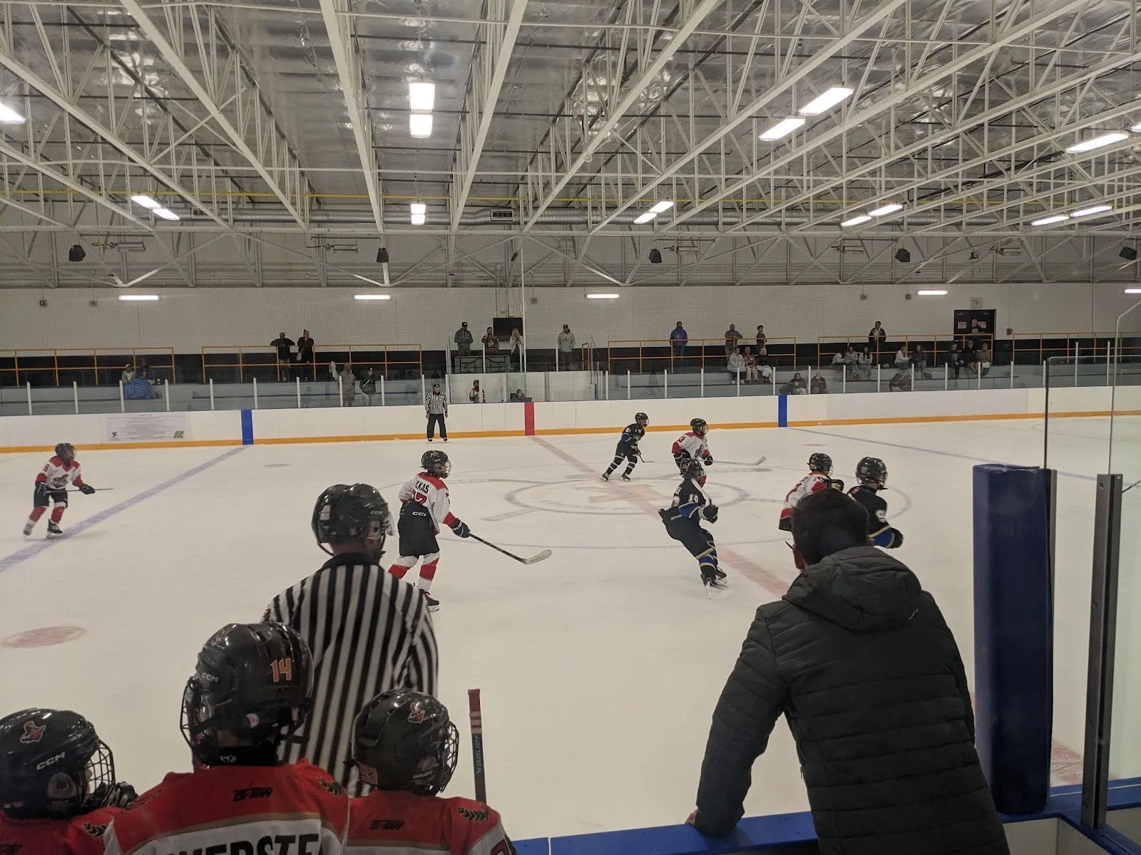 Una vista amplia de la pista vista a través del vidrio, con jóvenes jugadores repartidos por el hielo alrededor de una portería y una línea de vallas naranja bajo un techo claro. — Bill Burgoyne Memorial Arena