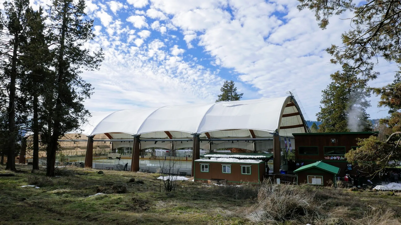 The arena's white tensioned roof seen from across a grass field on a sunny day, with pine trees in front and a small cabin below the roof. — Bill Collier Ice Arena