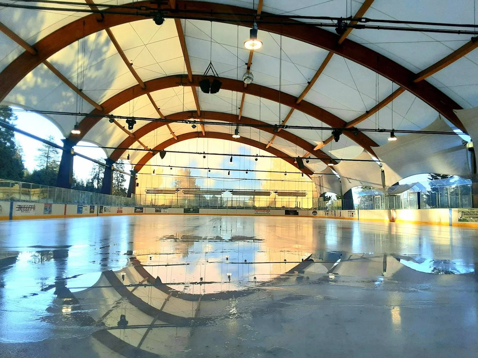 The ice sheet under the arena's open-sided tensioned roof with timber arches, empty ice reflecting the light, boards and hockey glass around it. — Bill Collier Ice Arena