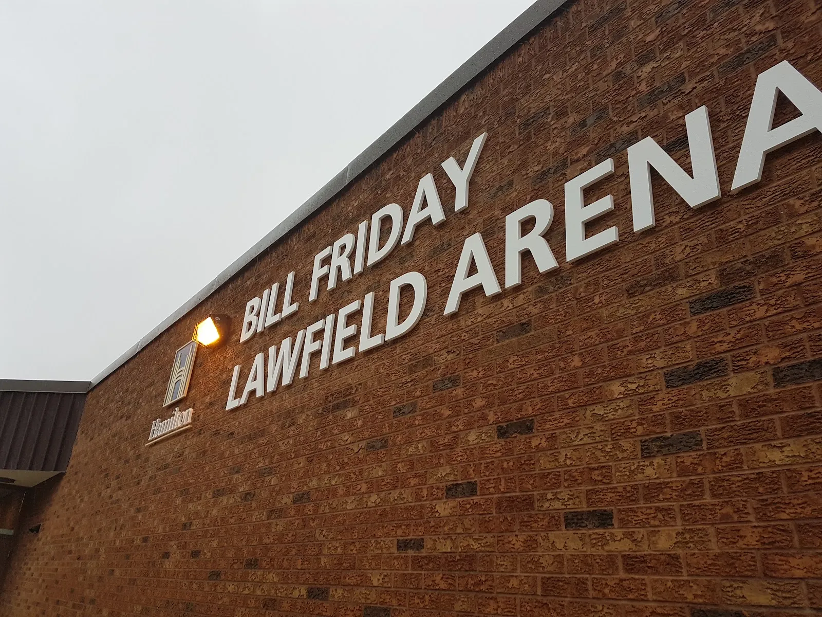 Muro de ladrillo de la arena con grandes letras que dicen "Bill Friday Lawfield Arena" y el logotipo de la ciudad de Hamilton. — Bill Friday Lawfield Arena