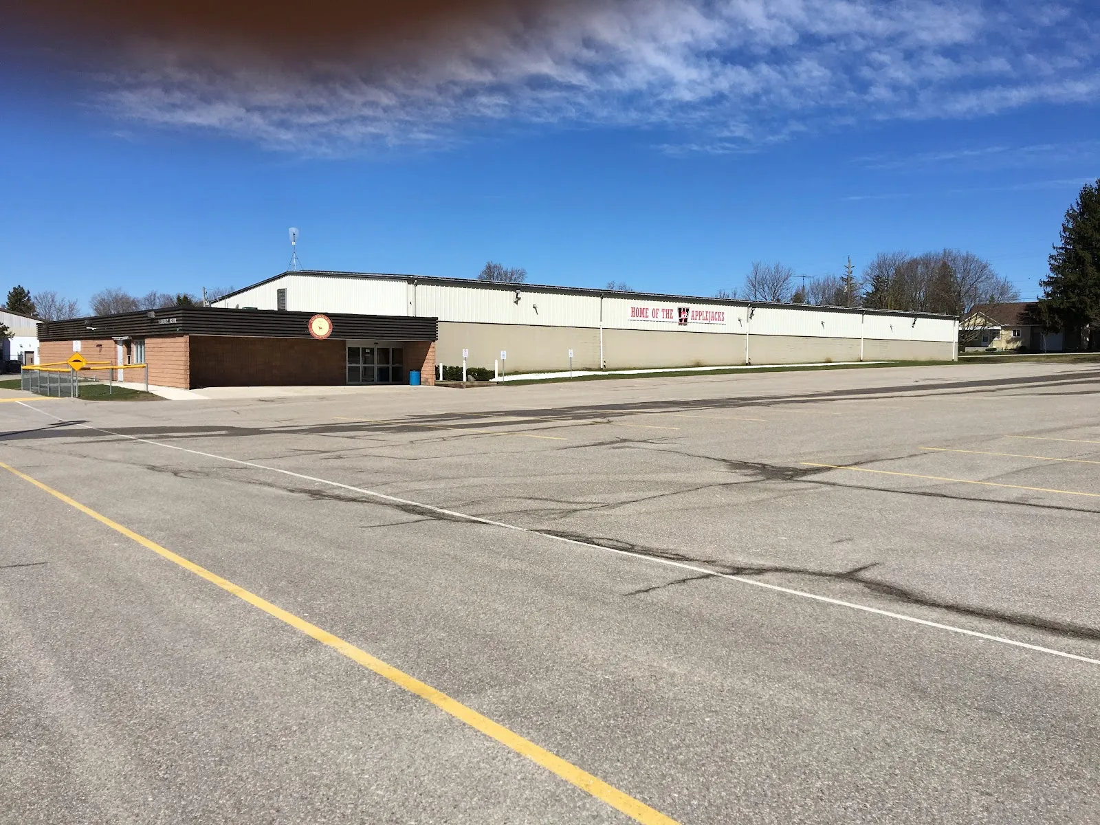 El largo y bajo edificio del Bill Gies Recreation Centre, con un ala de entrada de ladrillo y un bloque de pista revestido de metal blanco y gris, visto al otro lado de su aparcamiento de asfalto bajo un cielo azul. — Bill Gies Recreation Centre