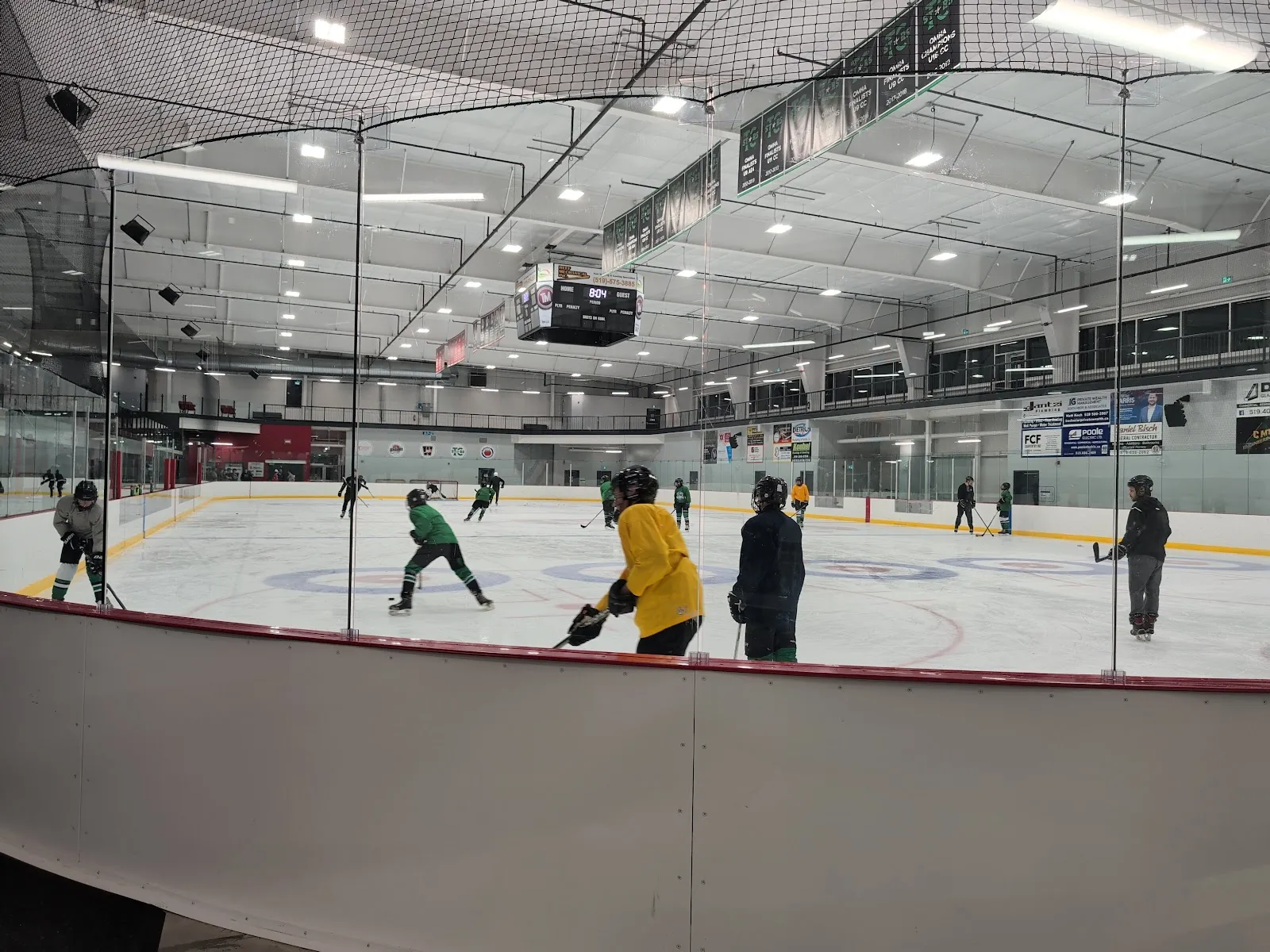 The ice from behind the boards during a skate, with a scoreboard, banners along the far wall and a steel-framed roof. — Bill Gies Recreation Centre