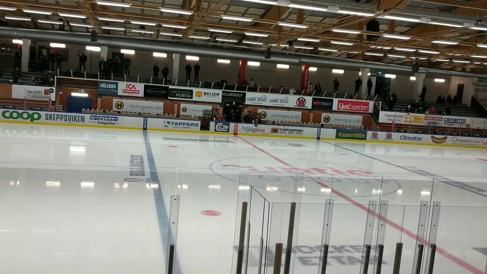 Empty ice at Billingehov in Skövde seen from the glass above the boards, with the blue line, centre circle and sponsor boards, under the timber roof, a few spectators far away in the stands — Billingehov ishallar