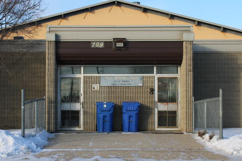 La entrada principal de la arena: muro de ladrillo color arena con el número 709 de la calle y el letrero de la arena sobre puertas de vidrio, dos contenedores azules de reciclaje al frente y nieve en el suelo. — Billy Mosienko Arena