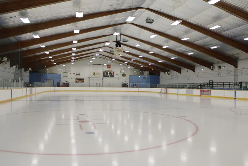 El hielo vacío desde el centro de la pista, con el círculo central rojo, las vallas y el cristal, y vigas de madera sobre paneles blancos en el techo. — Billy Mosienko Arena