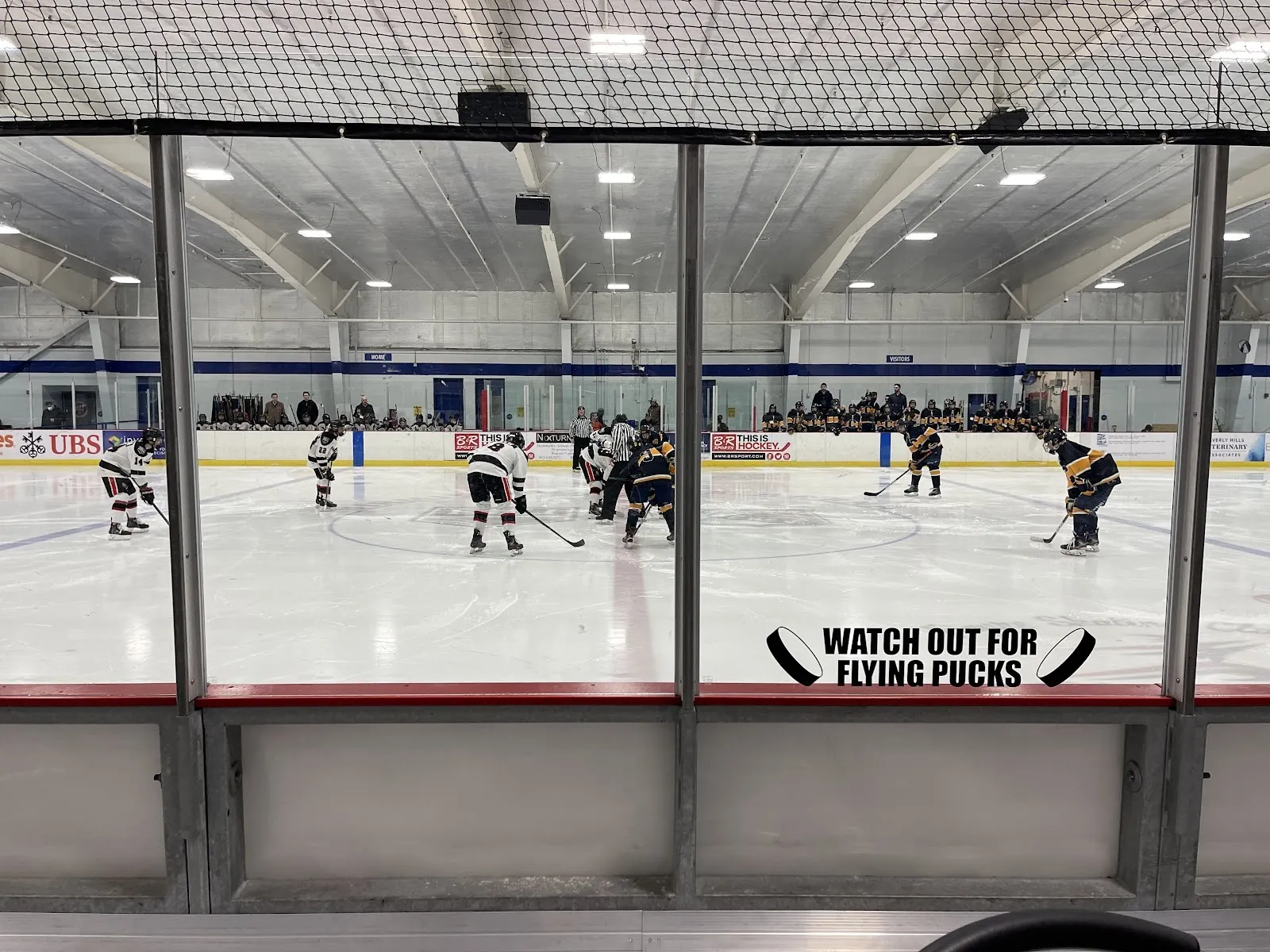A game seen through a glass panel at the boards, players spread across the wide sheet under a high steel roof, a Watch out for flying pucks sign on the boards. — Birmingham Ice Sports Arena