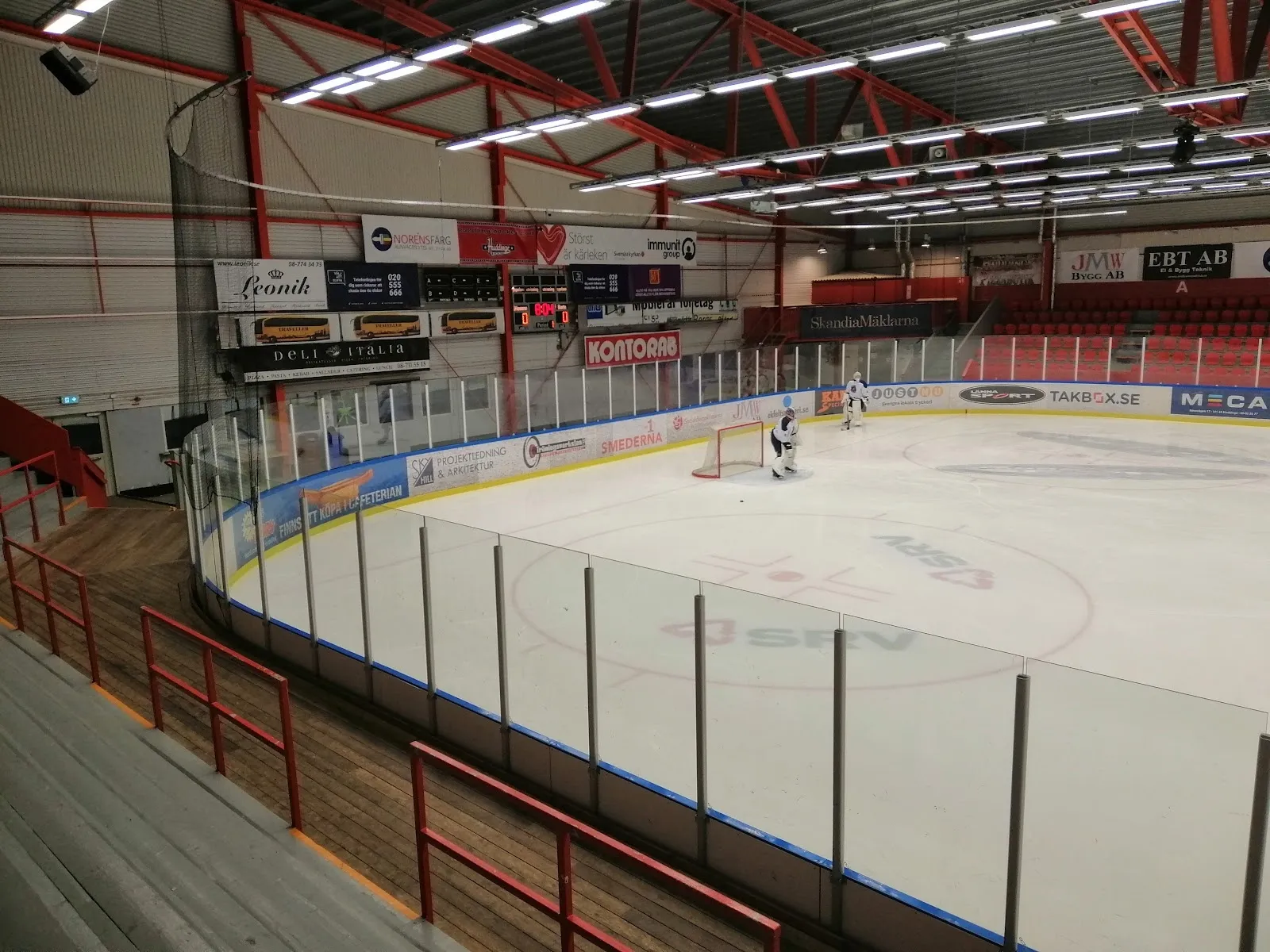 The ice of Björkängshallen B-hall in Huddinge from the side boards, with a few players on the ice, the red-trimmed side wall and the roof beams above. — Björkängshallen B-hall