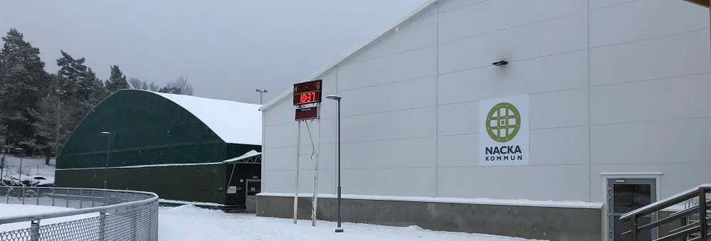 Björknäs ishalls vita vägg med Nacka kommuns logotyp, och det gröna istältet till vänster, i snön – Björknäs ishall