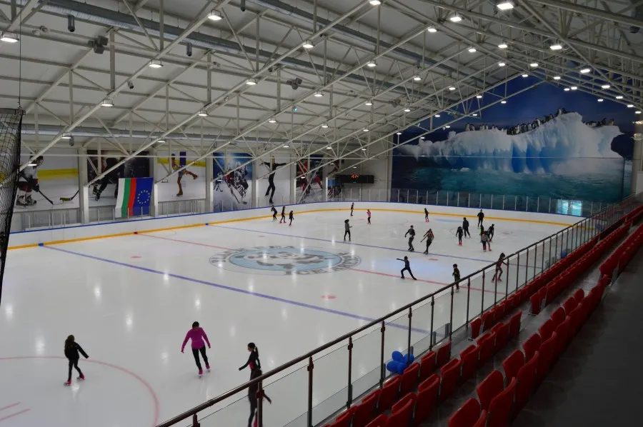 The full ice sheet of the arena seen from the stands, with skaters spread across the ice under a steel roof truss, a glacier mural on the end wall and red seating along the side. — Black Sea Ice Arena