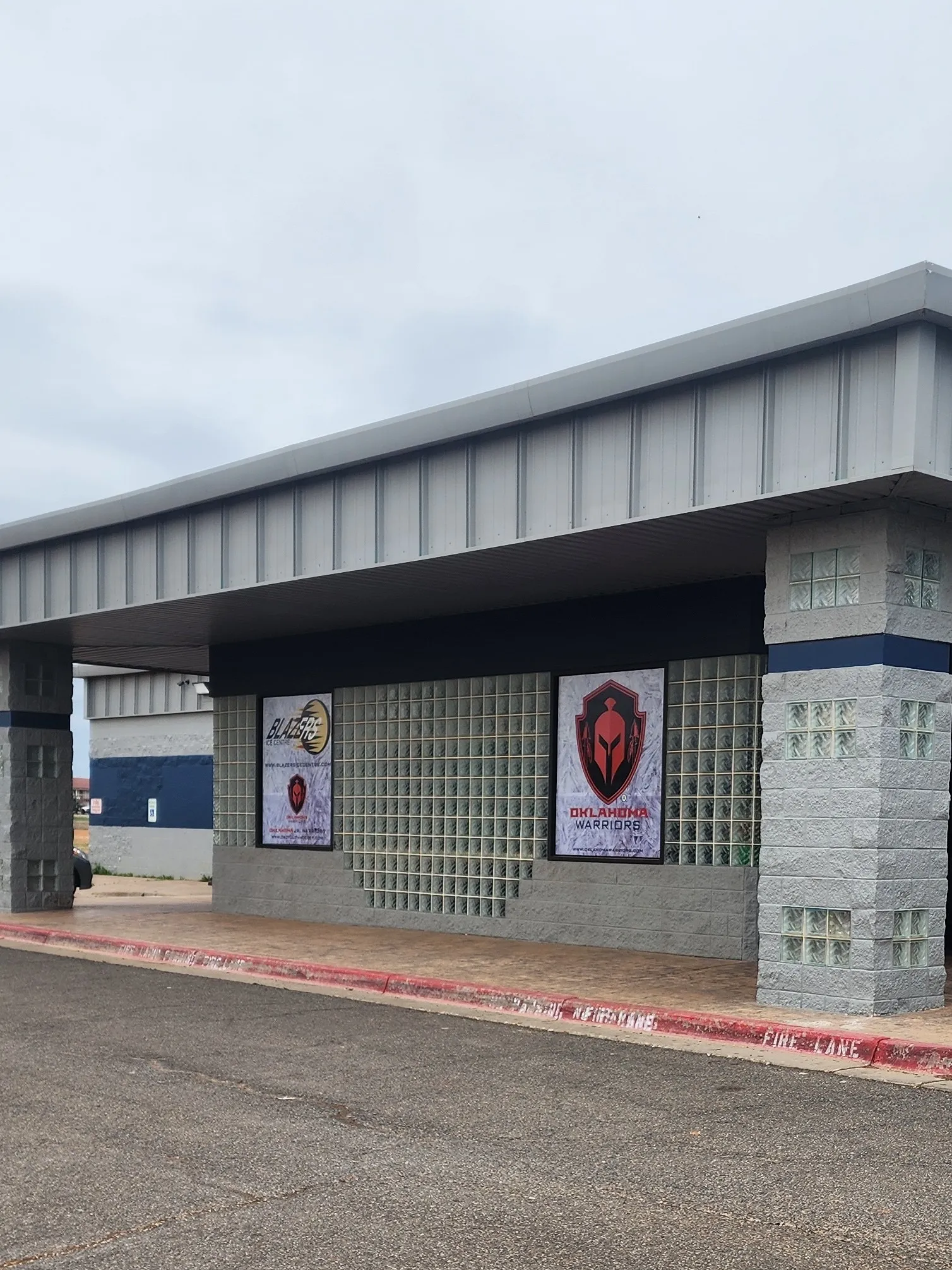 The Blazers Ice Centre entrance canopy over glass-block walls hung with Blazers and Oklahoma Warriors posters, a fire lane along the curb. — Blazers Ice Centre
