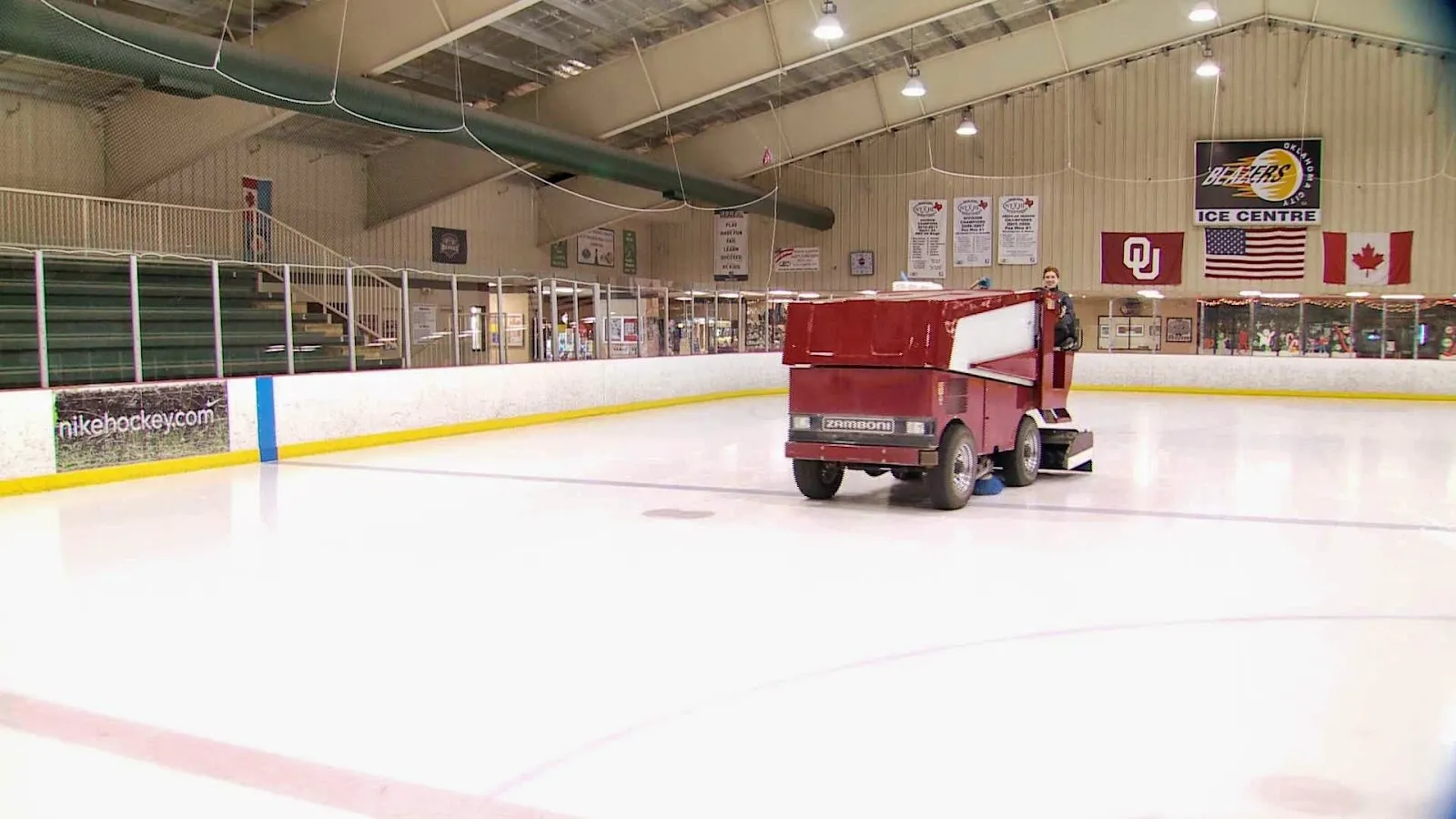 The resurfacer parked on a wide sheet, an Oklahoma Warriors banner and Canadian flag along the far wall under a timber and steel roof. — Blazers Ice Centre