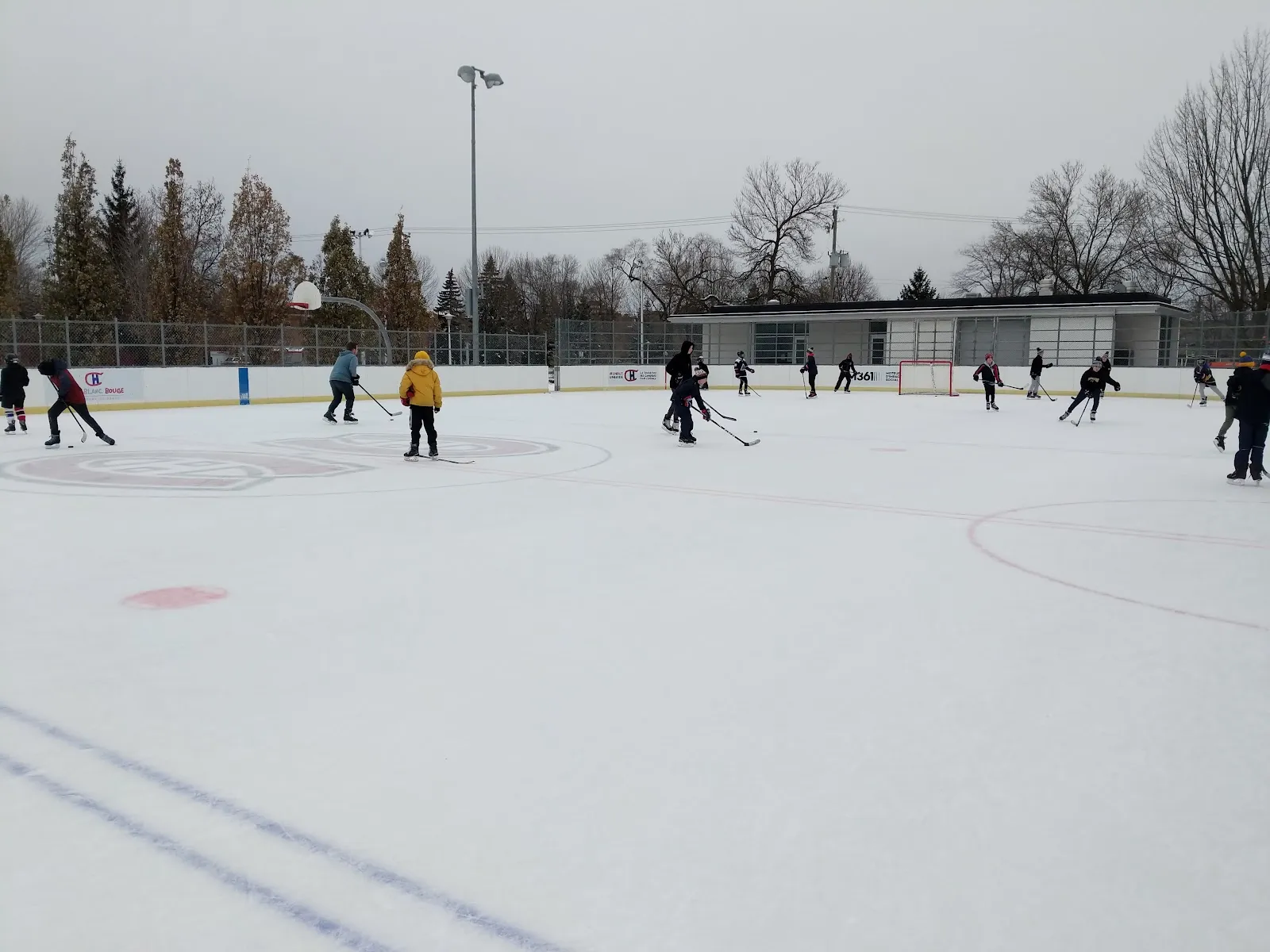 La patinoire réfrigérée Bleu Blanc Bouge du parc de la Confédération par temps couvert en hiver, des joueurs de hockey répartis sur la glace, un logo des Canadiens au centre et le chalet du parc en arrière-plan — Bleu Blanc Bouge Rink at Côte-des-Neiges–Notre-Dame-de-Grâce