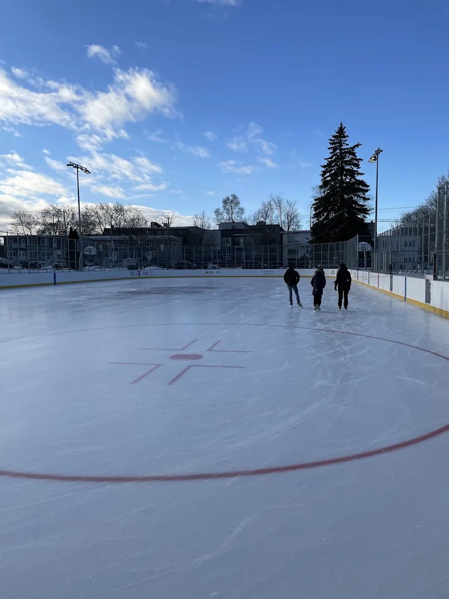 The refrigerated Bleu Blanc Bouge rink at Parc De Mésy in winter, a wide sheet of ice with a faceoff circle, three skaters seen from behind near the boards and a spruce tree and apartment buildings beyond — Bleu Blanc Bouge Rink at Parc De Mésy