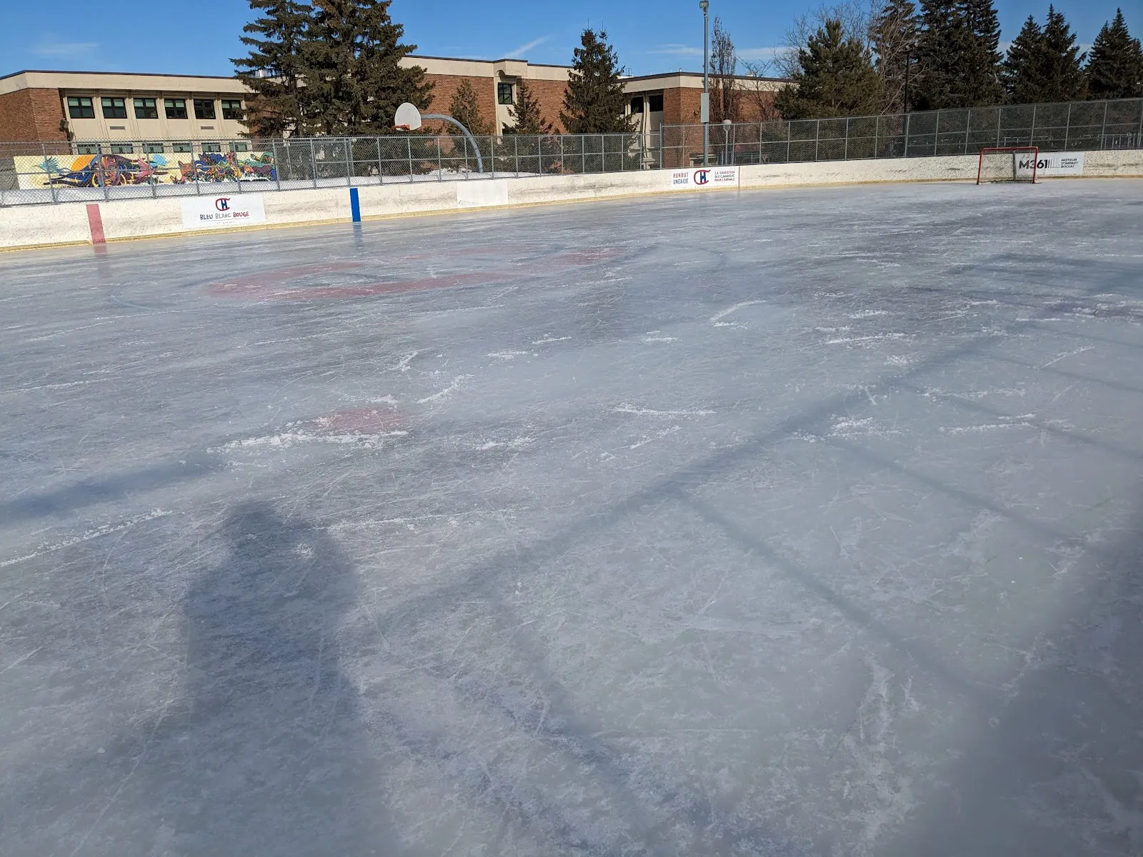 La patinoire réfrigérée Bleu Blanc Bouge du parc François-Perrault, vide, par une journée d'hiver ensoleillée : glace éraflée aux marques centrales à peine visibles, bandes avec panneaux de commanditaires, un but et des bâtiments scolaires en arrière-plan — Bleu Blanc Bouge Rink at Parc François-Perrault