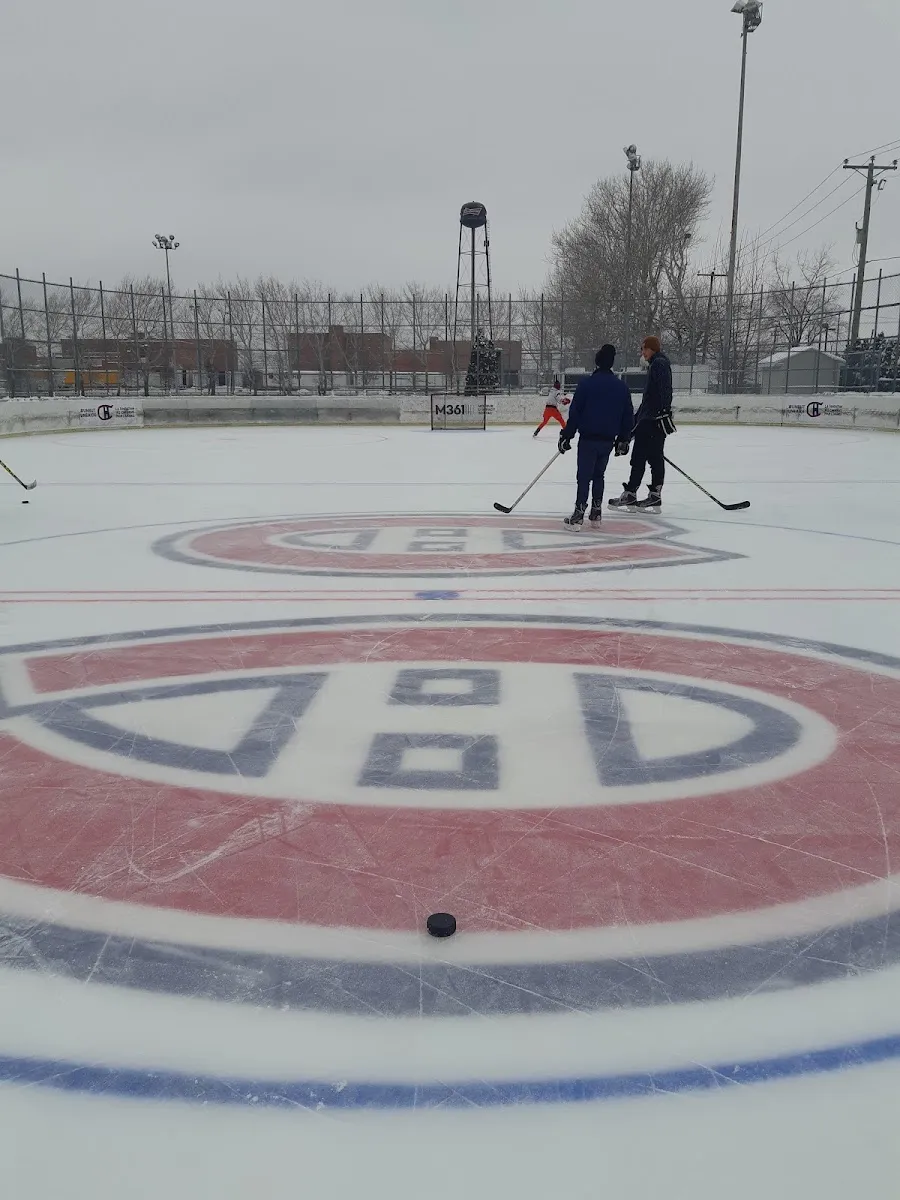La patinoire réfrigérée Bleu Blanc Bouge avec le logo des Canadiens peint au centre de la glace, une rondelle sur la glace, deux joueurs et un patineur au loin le long des bandes, une clôture et un château d'eau en arrière-plan — Bleu Blanc Bouge Rink at Parc Hayward