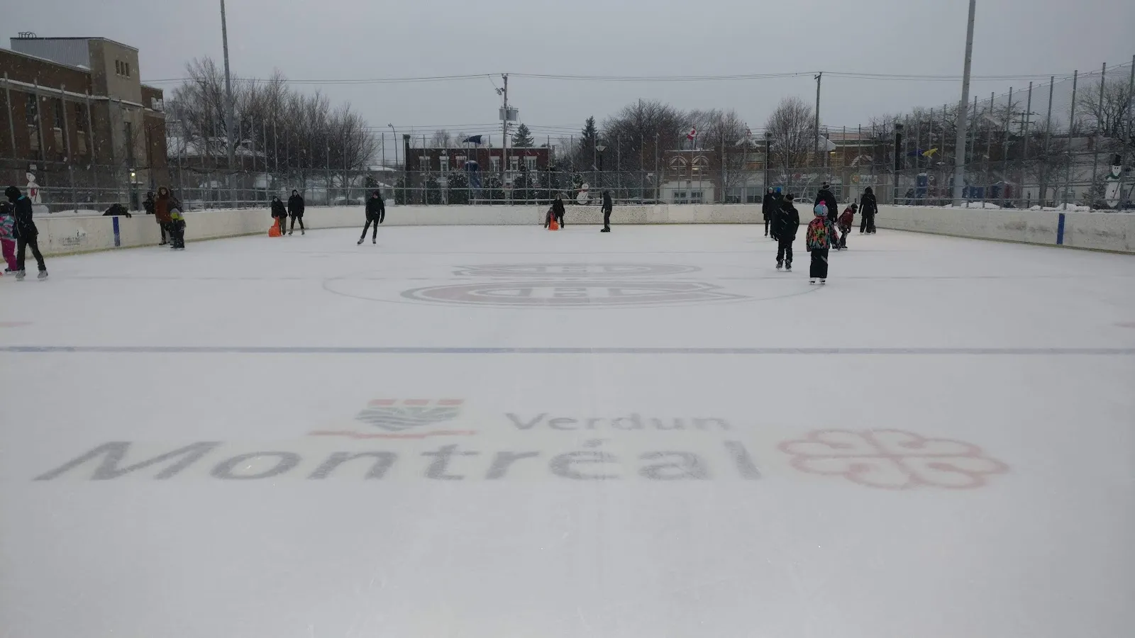 The Bleu Blanc Bouge rink at Parc Willibrord in Verdun with a Montréal and Verdun logo and a Canadiens logo painted on the ice, skaters near the boards and low buildings behind — Bleu Blanc Bouge Rink at Parc Willibrord