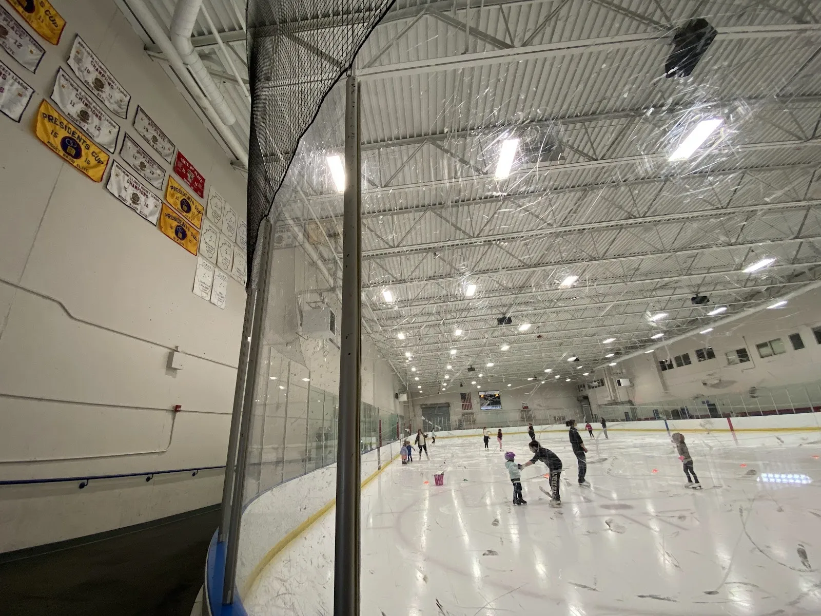 Un partido visto por encima del vidrio, con jugadores repartidos por una pista amplia bajo un alto techo de acero con un marcador en la pared izquierda. — Bloomington Ice Center