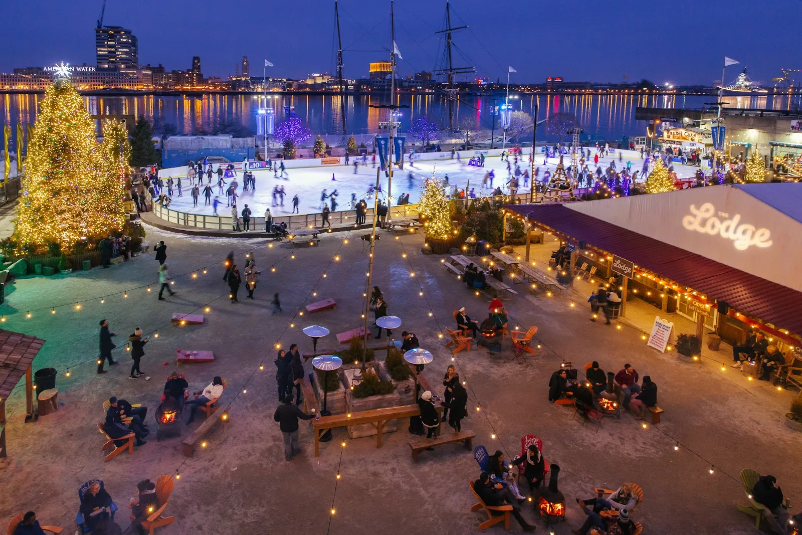 The rink at night from above, with the holiday tree, the Lodge and skaters on the ice against the Delaware River skyline. — Independence Blue Cross RiverRink Winterfest