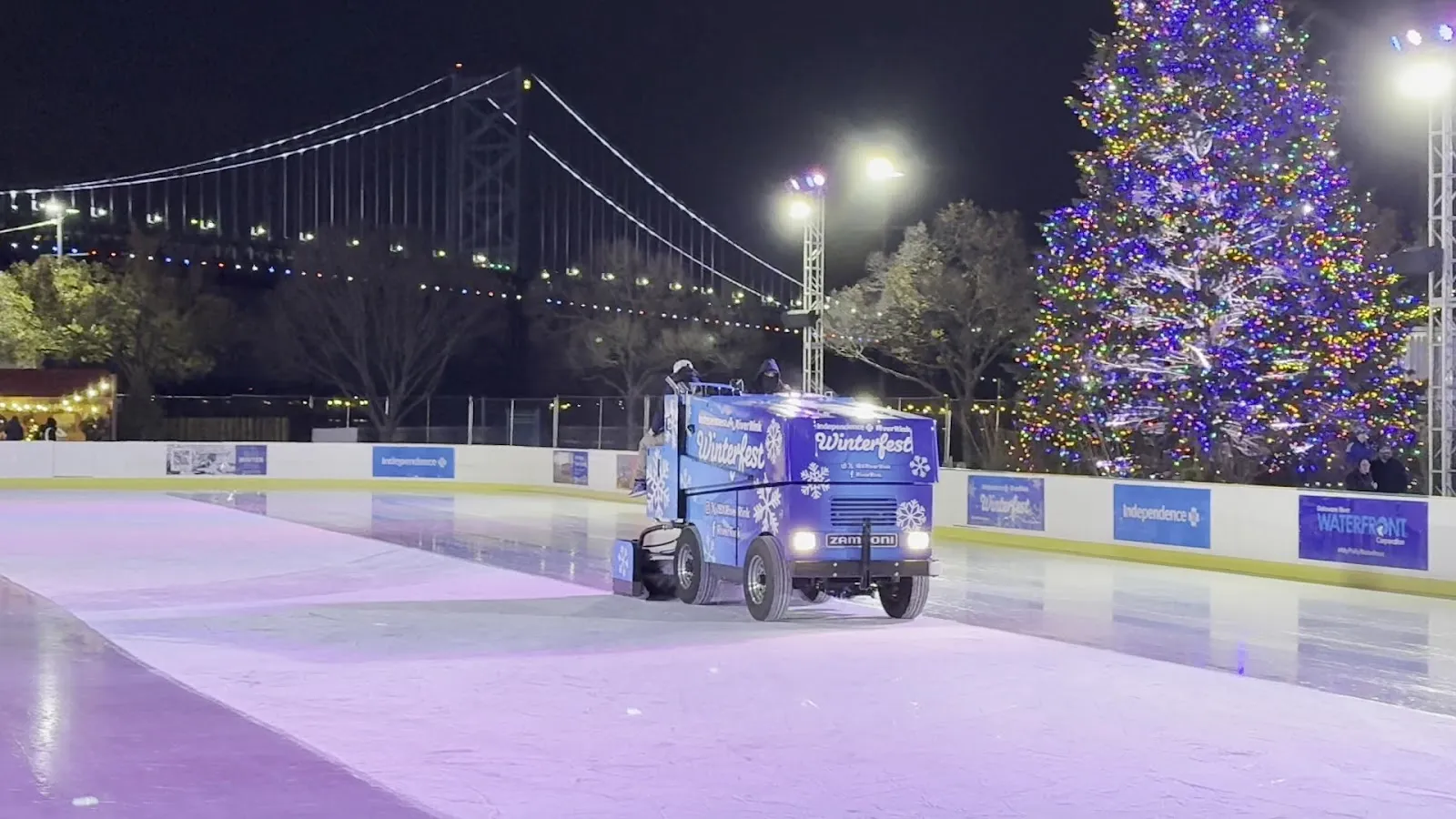 The Zamboni resurfacing the empty outdoor sheet at night, with the Ben Franklin Bridge and the holiday tree behind the boards. — Independence Blue Cross RiverRink Winterfest