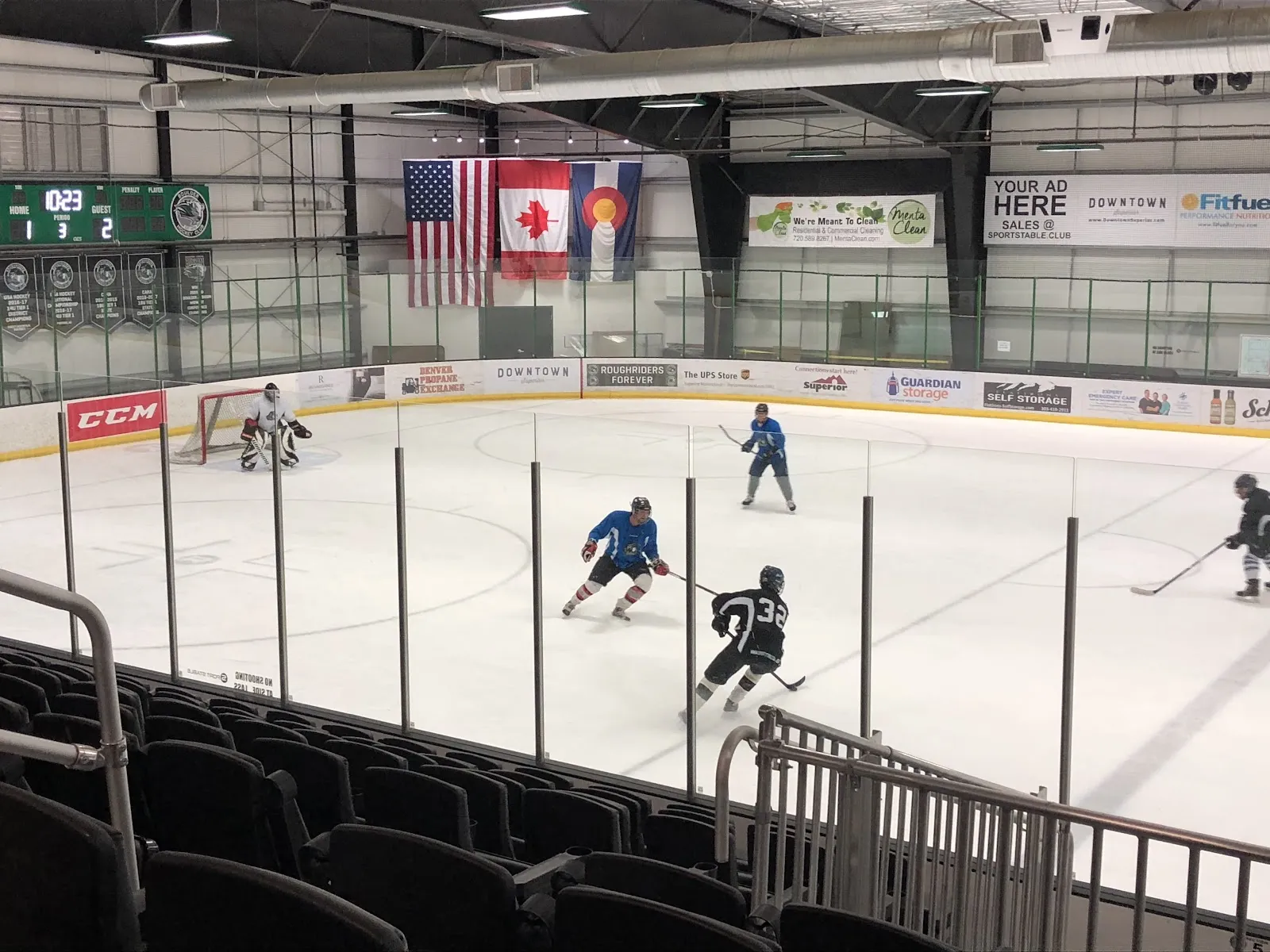 Un partido de hockey en una de las pistas visto desde las gradas, con banderas de EE. UU., Canadá y Colorado y un marcador detrás del cristal. — Blue Sport Stable