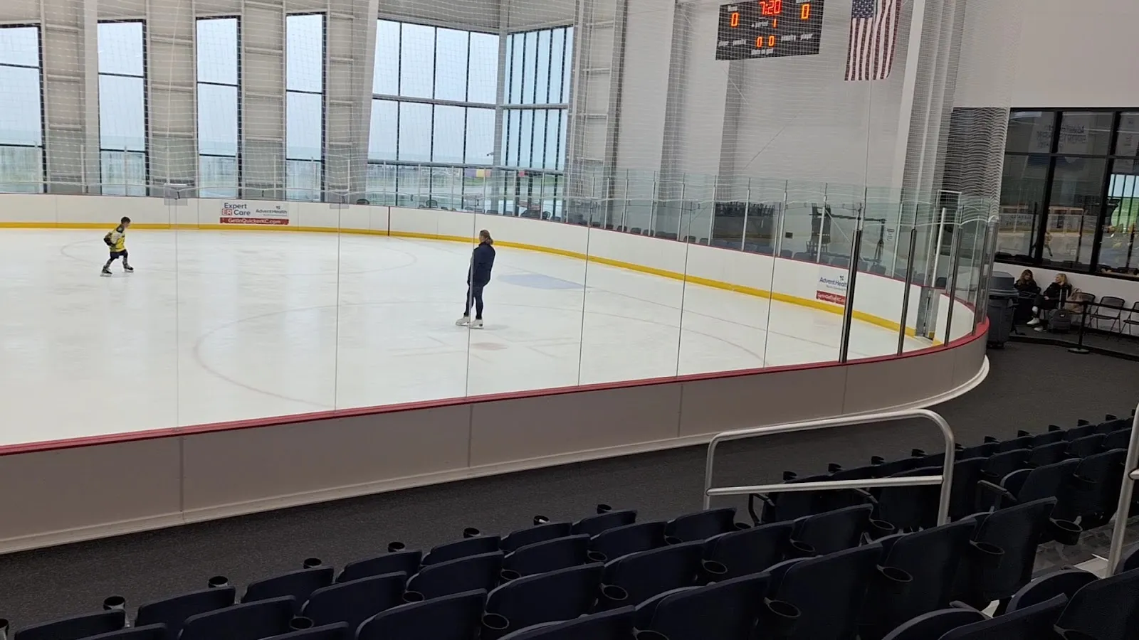 Two skaters on the ice seen from the upper seats past a curved glass rail, the bowl of the arena around the sheet and a tall window wall behind. — AdventHealth Sports Park at Bluhawk