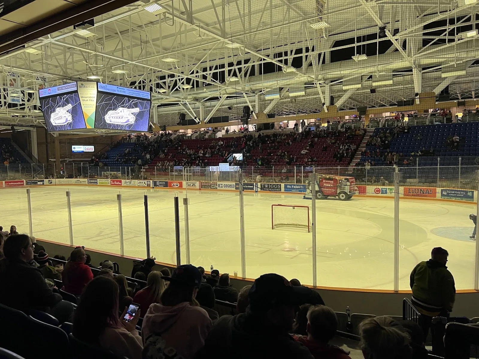 La arena vista desde las butacas bajas, con un partido de hockey en el extremo opuesto del hielo bajo un techo blanco de armaduras de acero con pantallas de video y espectadores en las gradas. — Boart Longyear Memorial Gardens