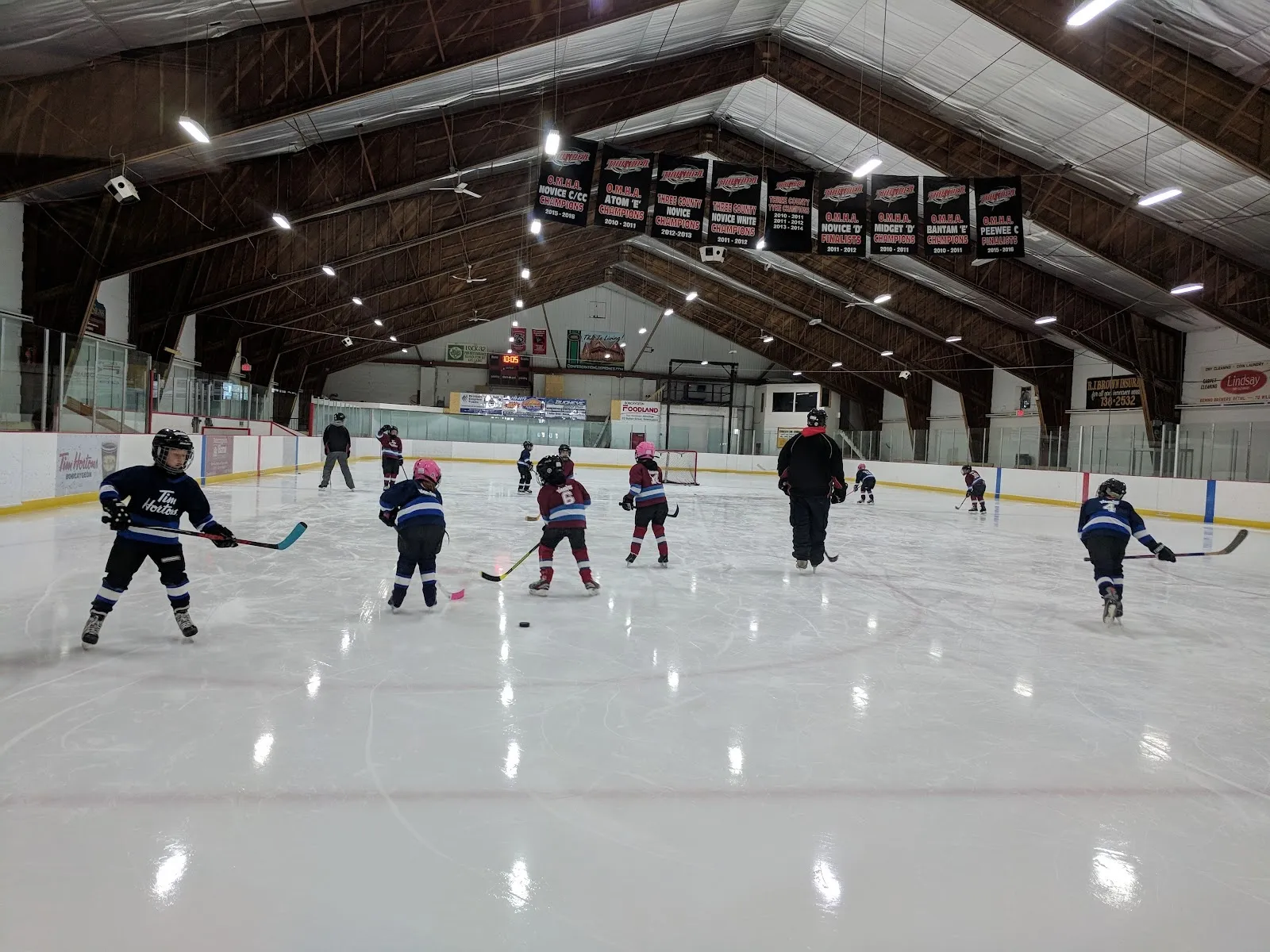 Young players lined up in a drill across a wide sheet under a dark timber roof hung with team banners, bright reflections from the lights on the ice. — Bobcaygeon/Verulam Community Centre