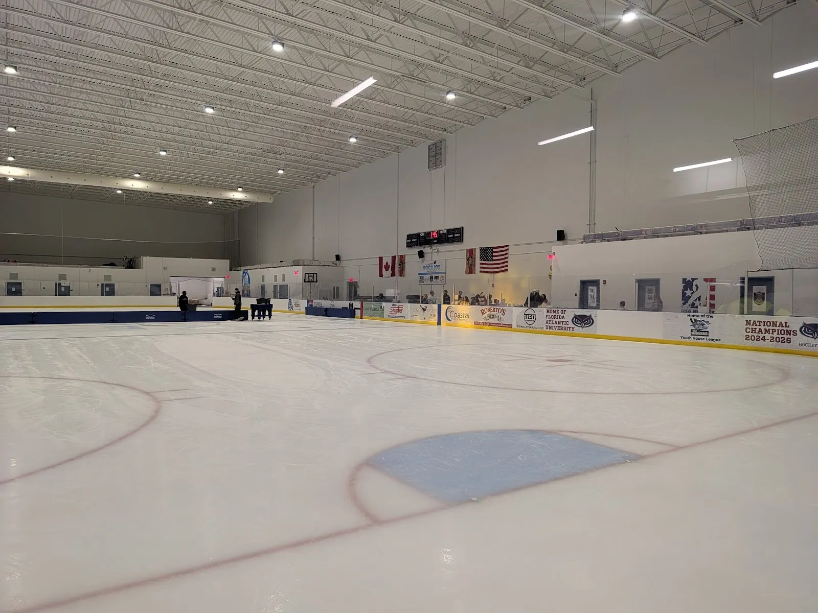 Ice-level view across a mostly empty NHL-size sheet toward the scoreboard, Canadian and U.S. flags and a "Home of Florida Atlantic University" dasher board, white steel roof trusses overhead; a few small figures by the far boards. — Boca Ice (Boca Raton)