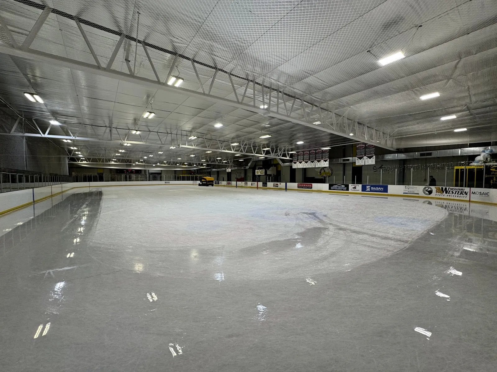 The empty sheet under the steel roof trusses and rows of lights, with sponsor boards along the dasher and a resurfacer parked at the far end. — Bode Ice Arena