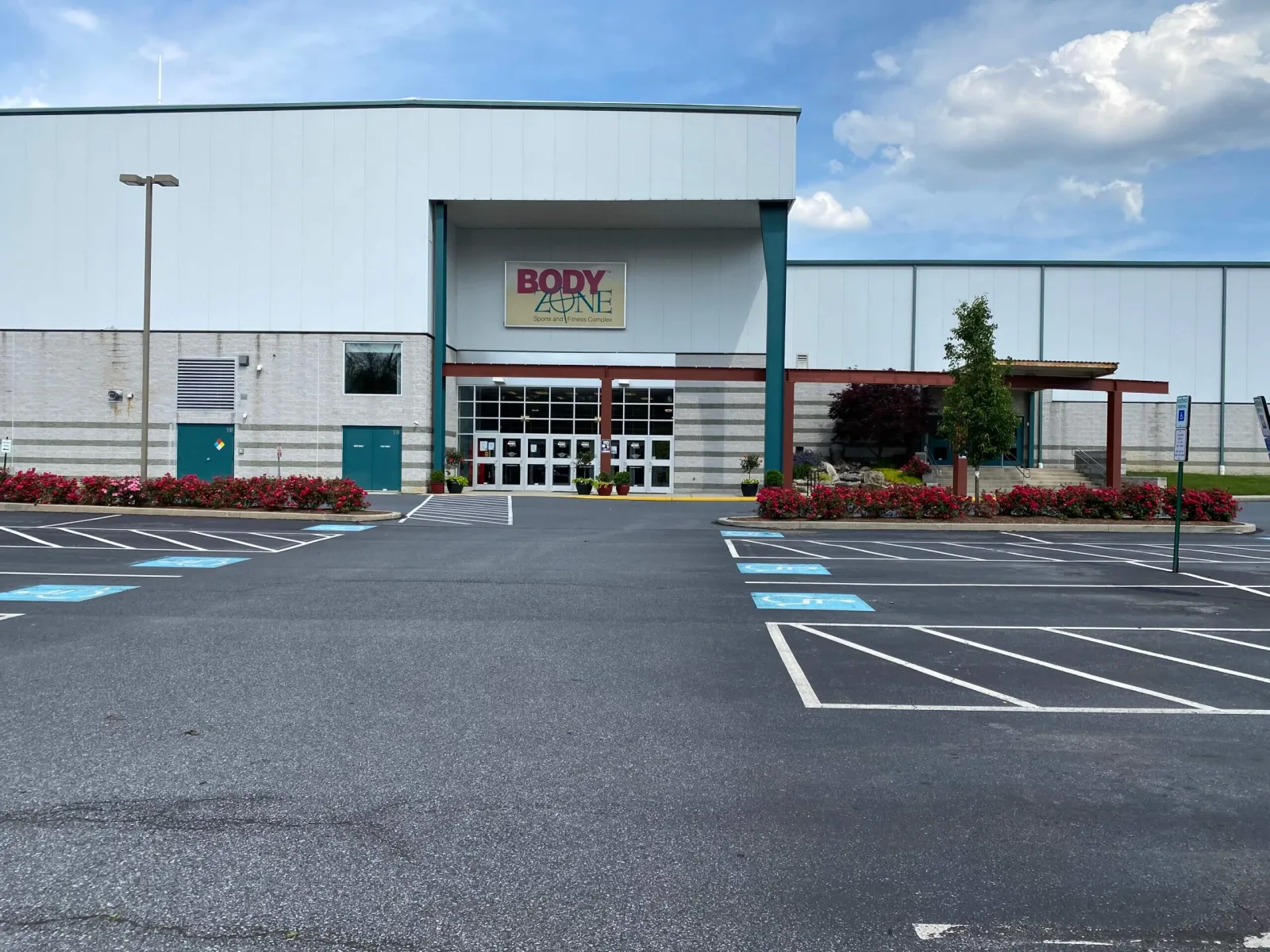 Front entrance of Body Zone Sports and Wellness Complex, a large steel-sided building with a BODY ZONE Sports and Fitness Complex sign over the glass doors, empty parking lot in front. — Body Zone Sports and Wellness Complex