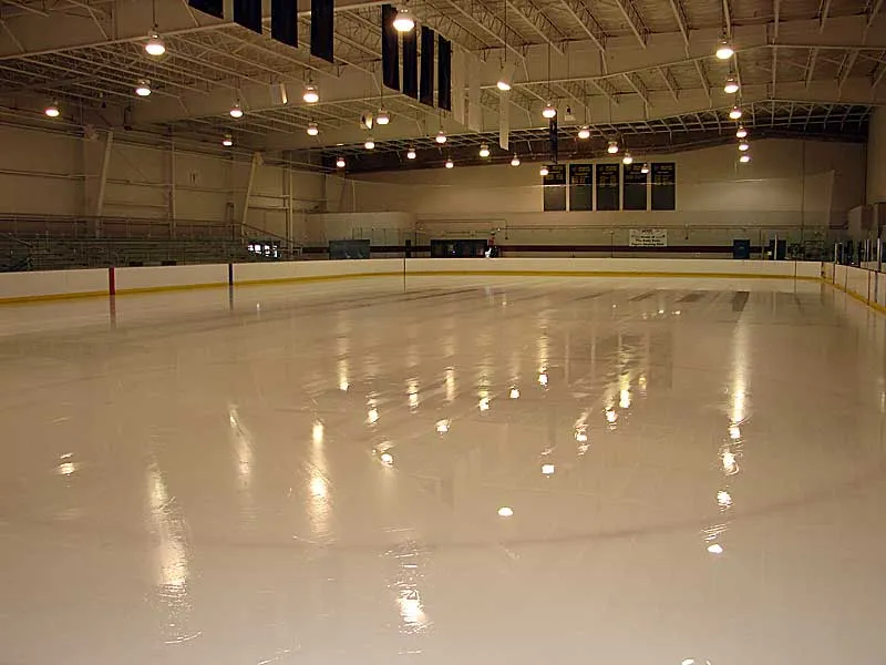Empty ice sheet at Body Zone with white dasher boards, bleacher seating on one side, and a scoreboard along the far wall. — Body Zone Sports and Wellness Complex