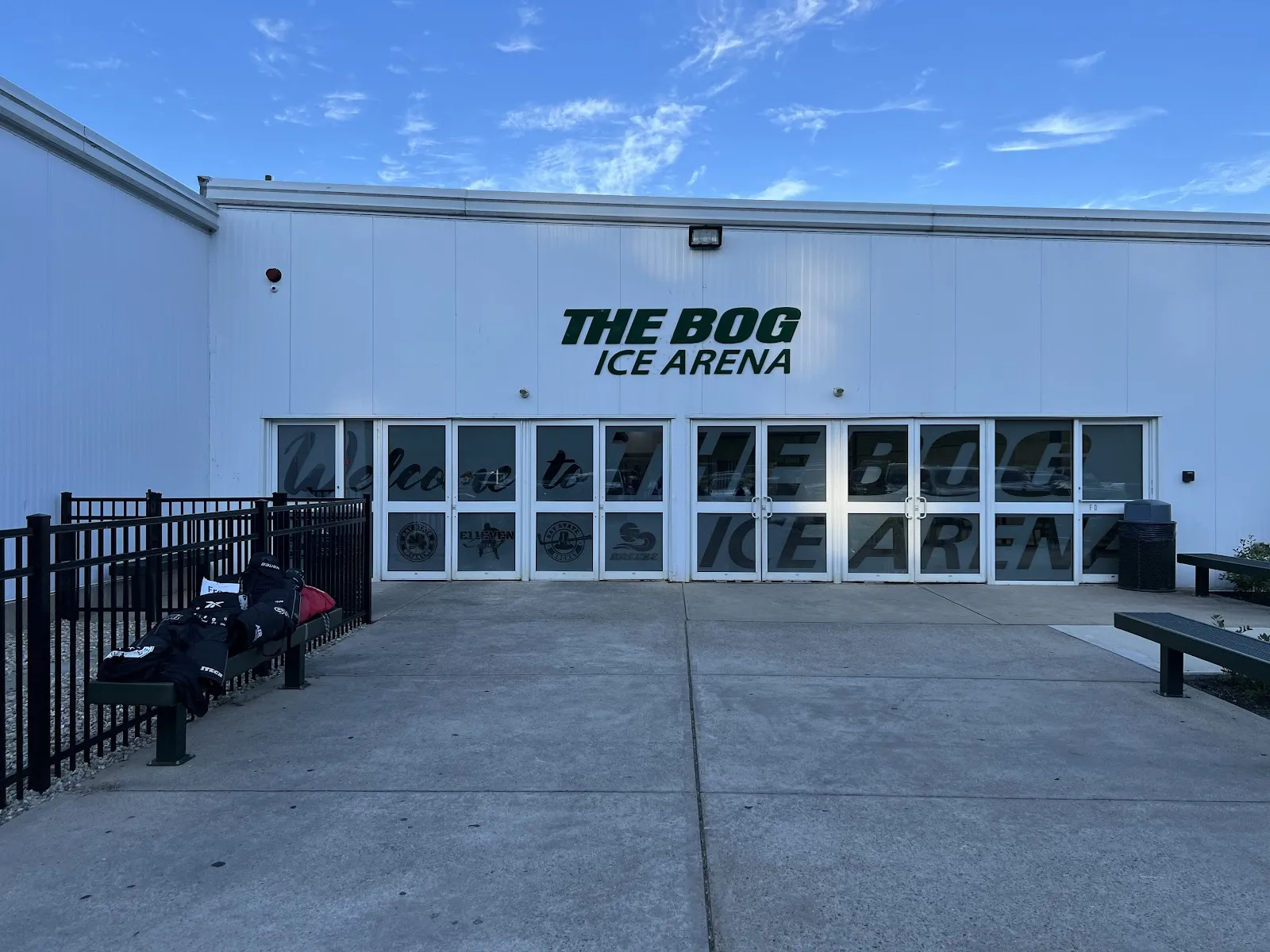 The white front of the arena with green "The Bog Ice Arena" lettering above a row of glass entry doors, a fenced walkway and benches out front. — The Bog Ice Arena