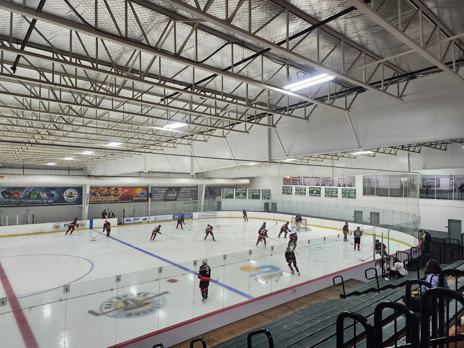 A wide view of the sheet from the glass, players spread across the ice around a centre logo under a steel-trussed ceiling with banners on the far wall. — The Bog Ice Arena