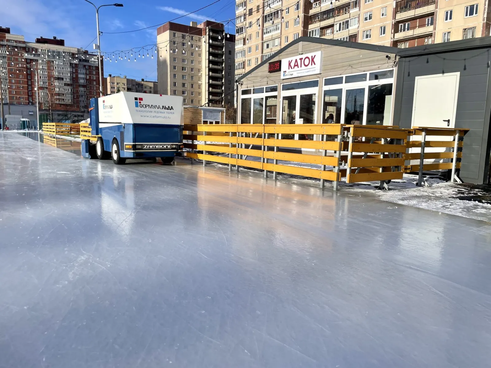 The Bogatyr open-air rink in Saint Petersburg by day: smooth ice, a Zamboni at the far side and the low wooden-fenced rink building marked KATOK in front of apartment blocks — Bogatyr Ice Rink Saint Petersburg