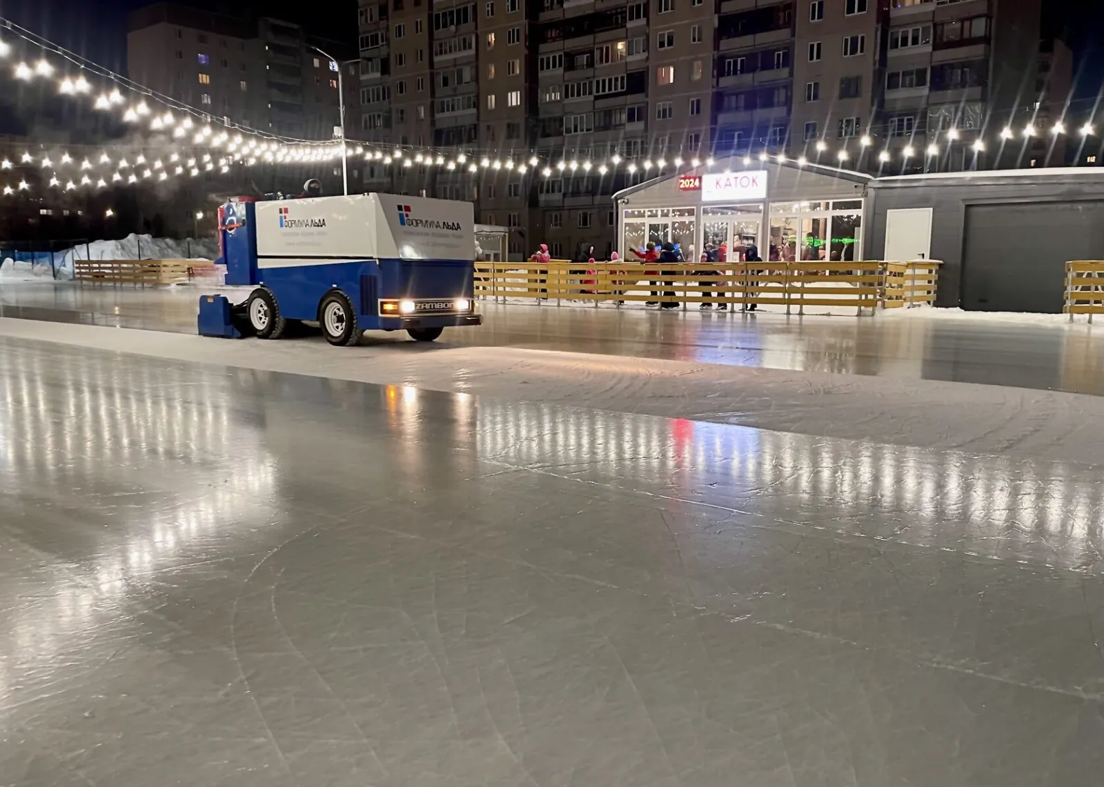 The empty ice of the Bogatyr rink at night, lit by strings of lights, with the resurfacing machine and the rink buildings along the far side — Bogatyr Ice Rink Saint Petersburg