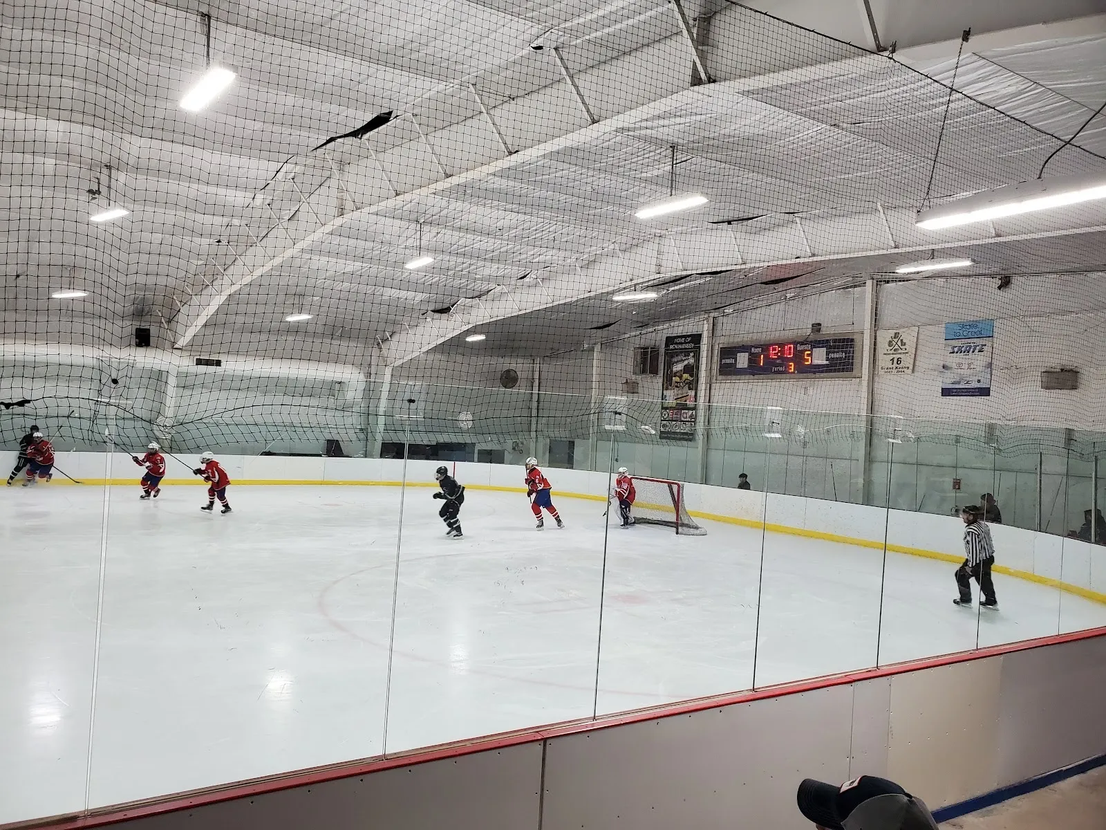 A game seen over the glass at the boards, players spread across the ice under an arched steel roof with sponsor banners along the far wall. — Bolton Ice Palace