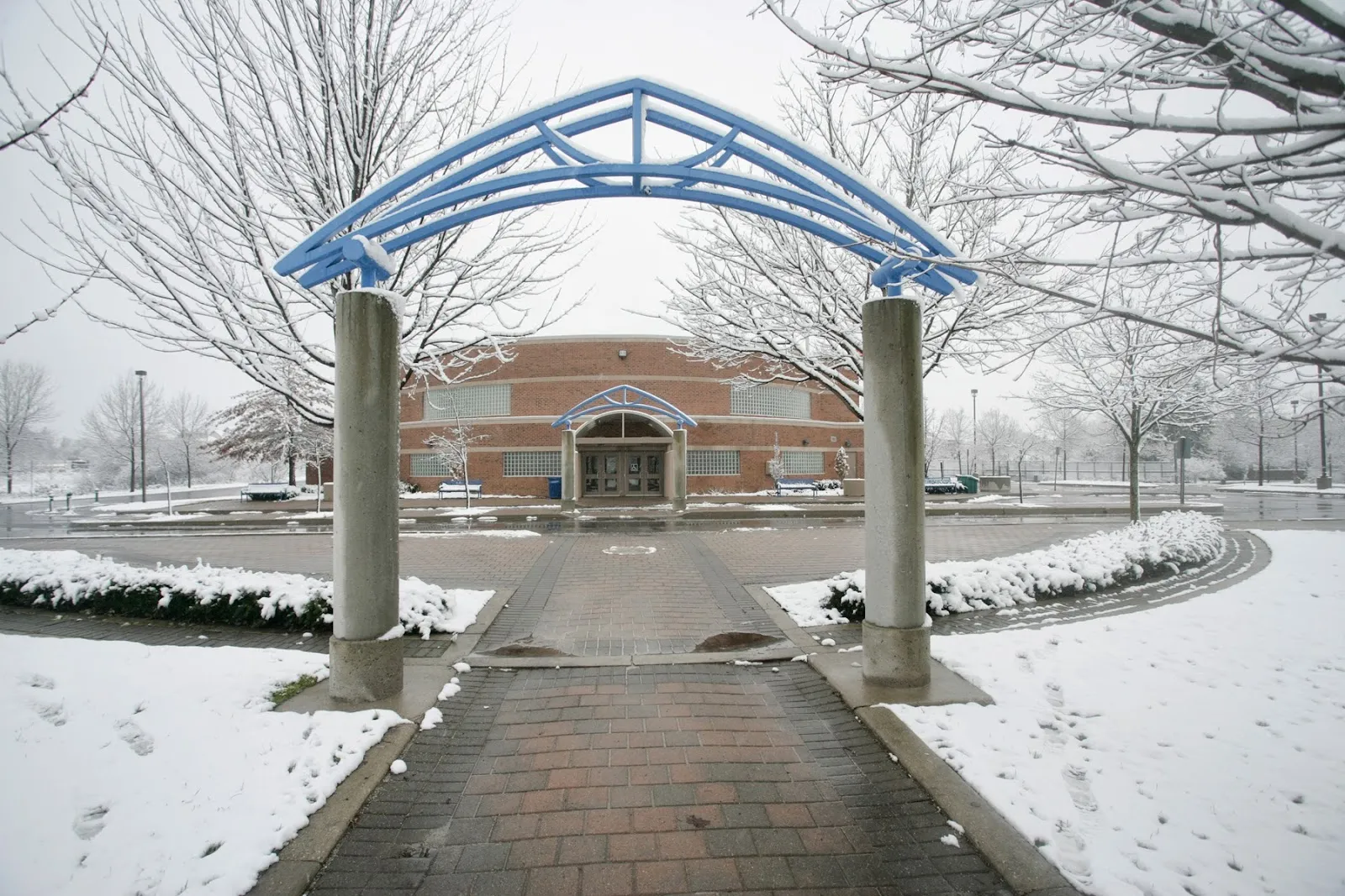 Un sendero nevado que pasa bajo un arco de acero azul y lleva a la entrada de ladrillo de la arena. — Bond Lake Arena (Richmond Hill)