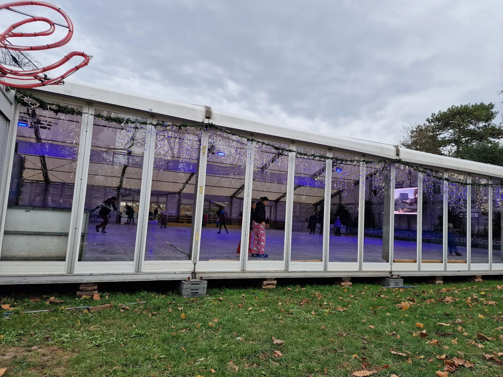 The long glazed tent that houses the rink, seen from the lawn of the Stadtgarten with autumn leaves on the grass and the lit rink visible through the panels. — bonnonIce