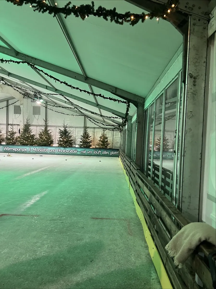 The empty ice lane inside the tent, lit in green, with fir trees painted along the boards and a garland running down the white canopy above. — bonnonIce