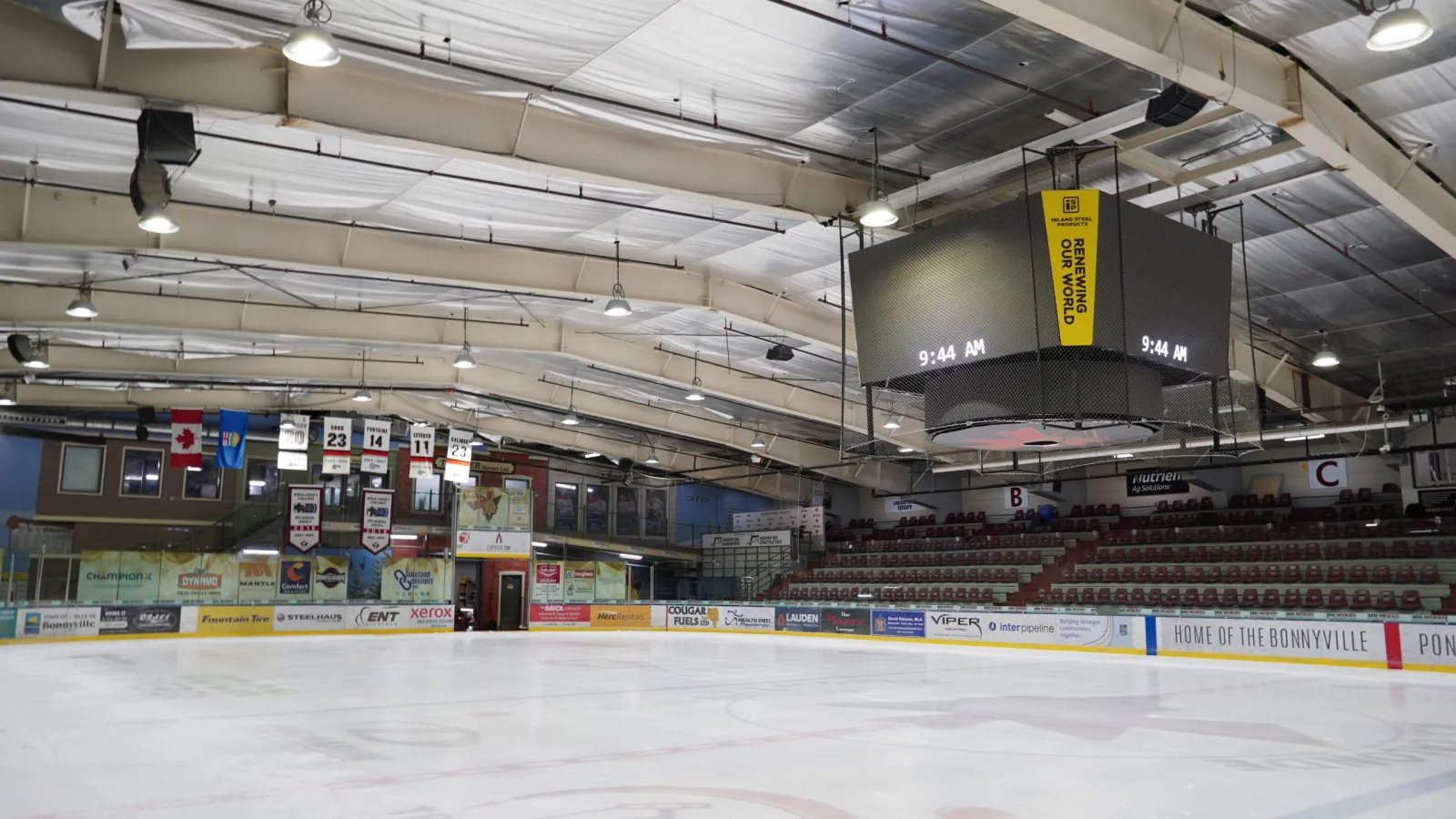 El hielo vacío de la R.J. Lalonde Arena bajo un marcador central colgante, con asientos rojos detrás de bardas que dicen Home of the Bonnyville Pontiacs. — Bonnyville & District Centennial Centre