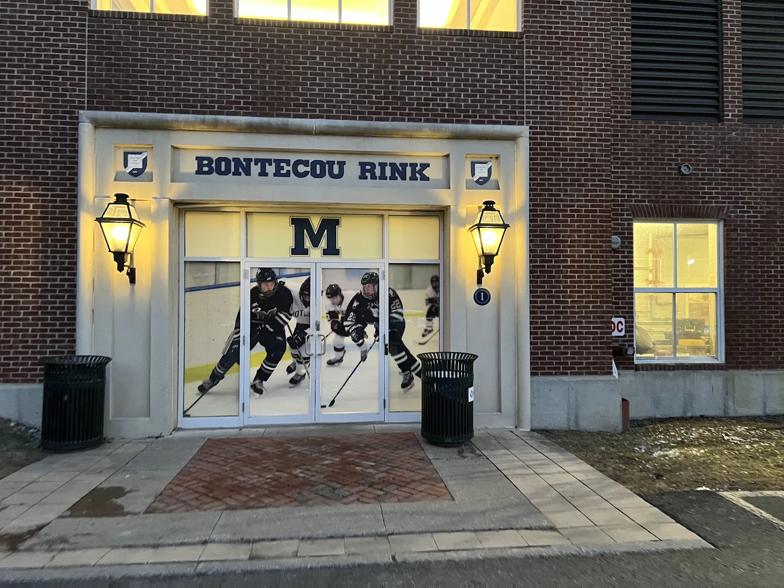 The brick Bontecou Rink entrance with its lit lanterns and arched doorway, hockey players visible through the glass. — Bontecou Rink at Millbrook School