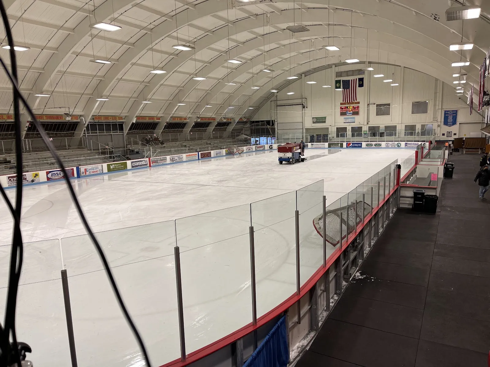 The empty rink under the arched white roof, seen from the boards, with an ice resurfacer at the far end beneath an American flag. — B.O.R. Arena