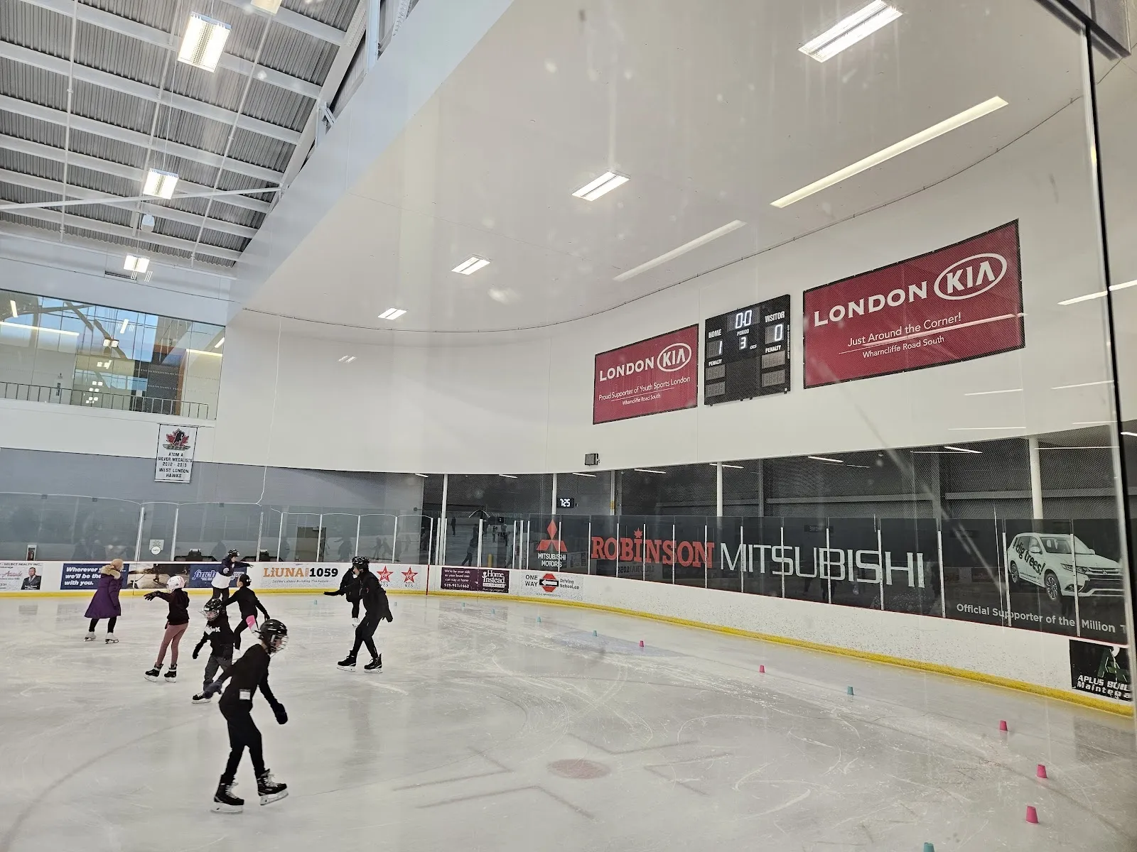 Patinadores del público en la superficie de hielo del Bostwick Arena, con la pared curva de vidrio blanco, banderines de patrocinadores y un marcador sobre las vallas y el pasillo superior detrás. — Bostwick Arena