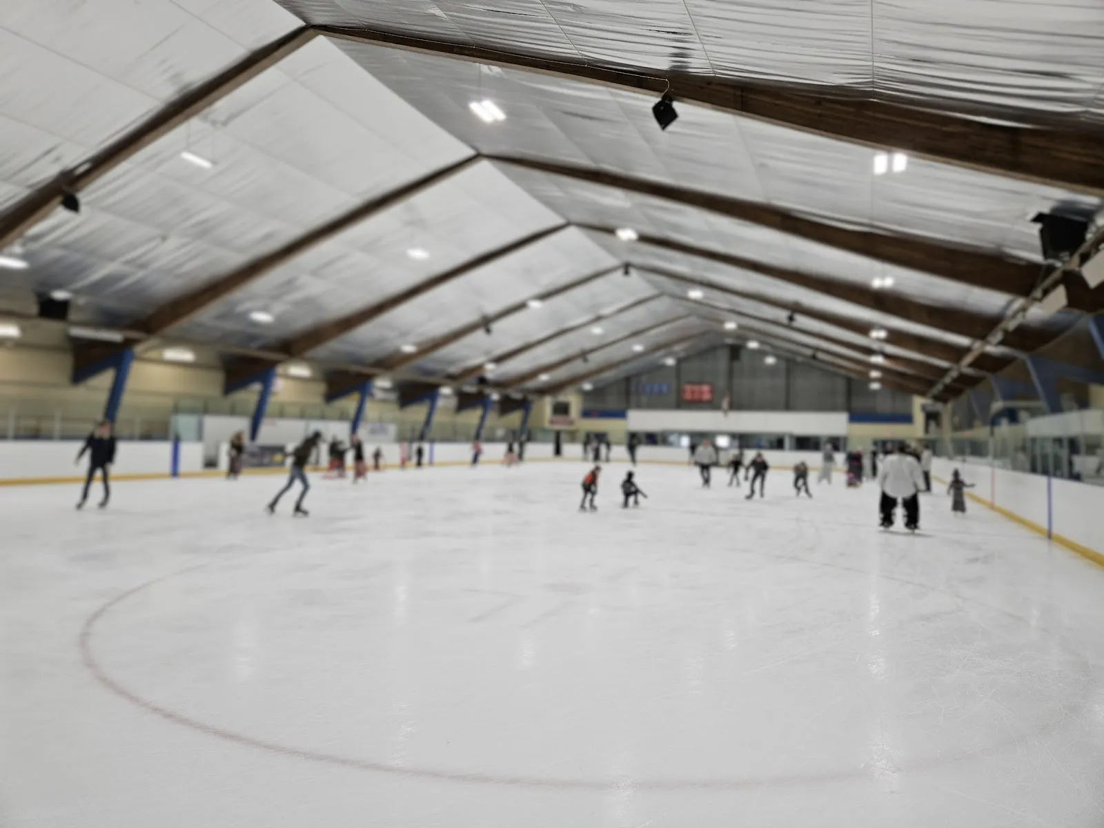 Patinadores repartidos por una pista amplia vistos desde las gradas, con un largo techo de armaduras de acero arriba y una brillante superficie de hielo blanco. — Bothwell Arena