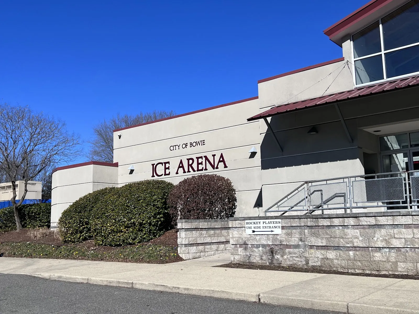 La fachada de la arena con las letras 'City of Bowie Ice Arena' en el muro de estuco, una entrada de techo rojo con escaleras y arbustos podados. — Bowie Ice Arena