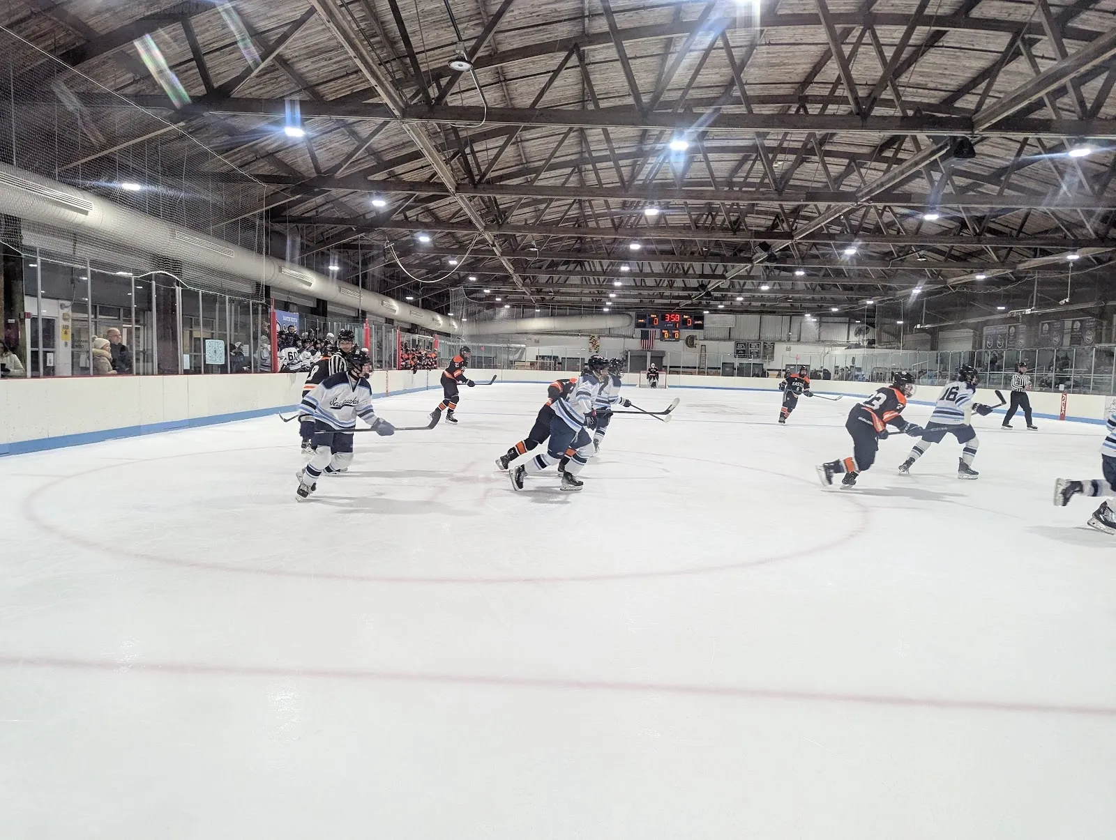 Una pista amplia vista desde las gradas, con jugadores repartidos por el hielo bajo un oscuro techo de armaduras de acero con banderines y anuncios de patrocinadores en la pared del fondo. — Bowie Ice Arena