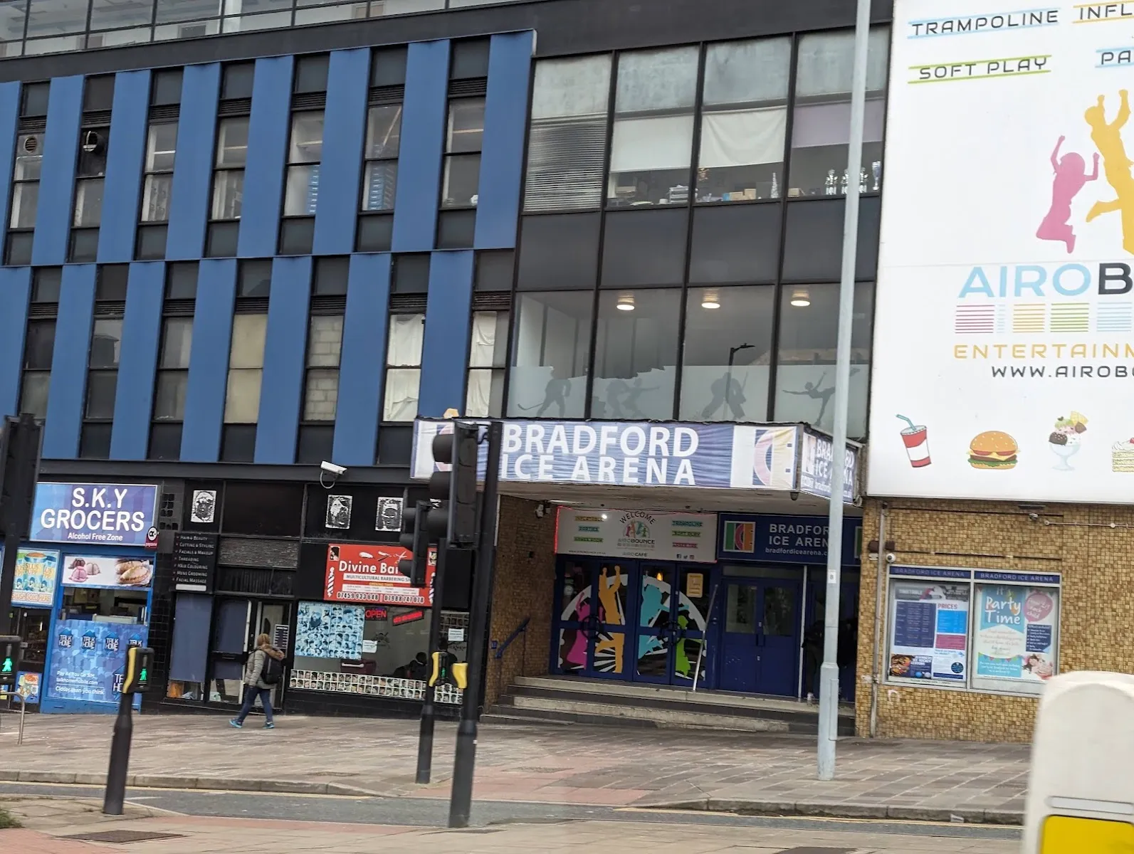 The street frontage of Bradford Ice Arena on Little Horton Lane: a blue 'Bradford Ice Arena' sign over a pillared entrance, with a grocer's shop beside it and a soft-play banner on the upper wall. — Bradford Ice Arena