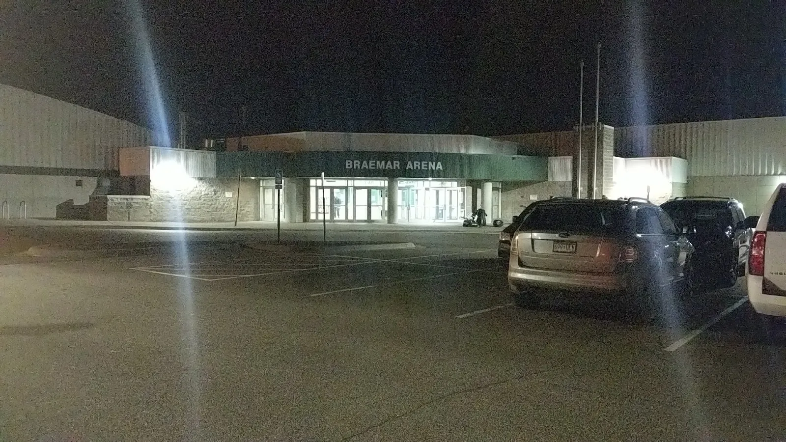 La entrada con techo verde de Braemar Arena con su nombre sobre las puertas de vidrio, iluminada de noche al otro lado del estacionamiento — Braemar Arena