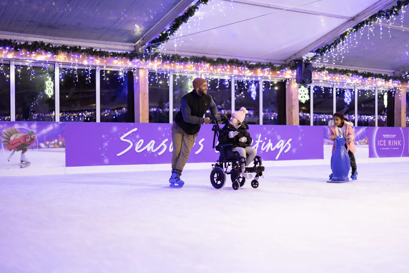 Skaters on the covered, blue-lit Braintree Village rink, one pushing a wheelchair across the ice, with boards and festive lights along the tent walls. — Braintree Village Ice Rink