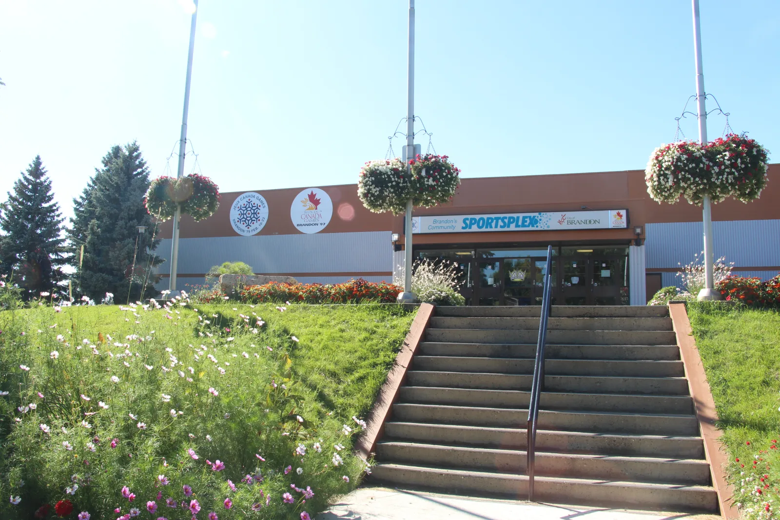Front entrance of the Sportsplex on a sunny day, with the "Brandon's Community SPORTSPLEX / City of Brandon" sign over the doors and hanging flower baskets on the flagpoles. — Brandon Community Sportsplex